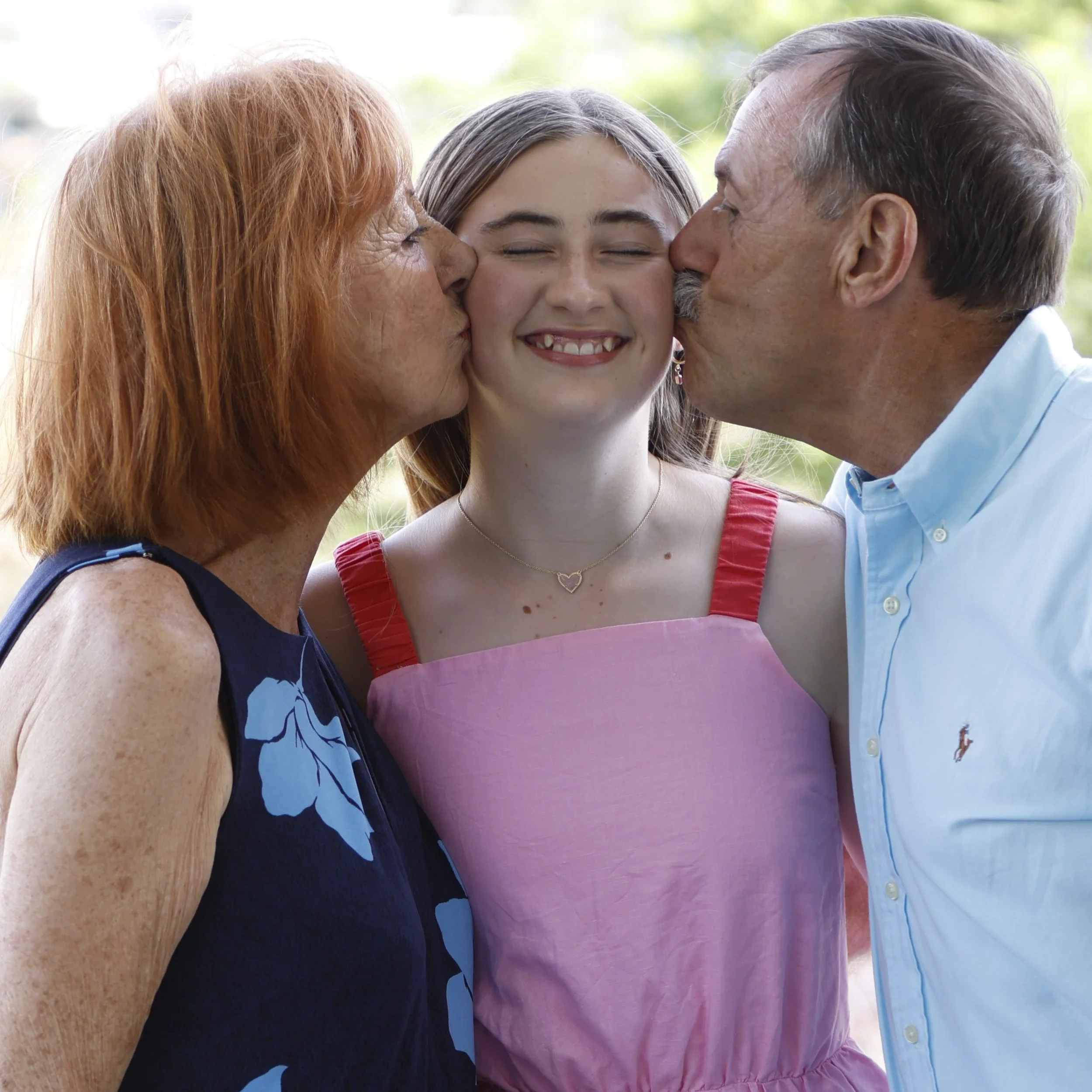 A young woman with disability, smiling with eyes closed, receiving kisses on each cheek from her parents, outdoors with greenery in the background.