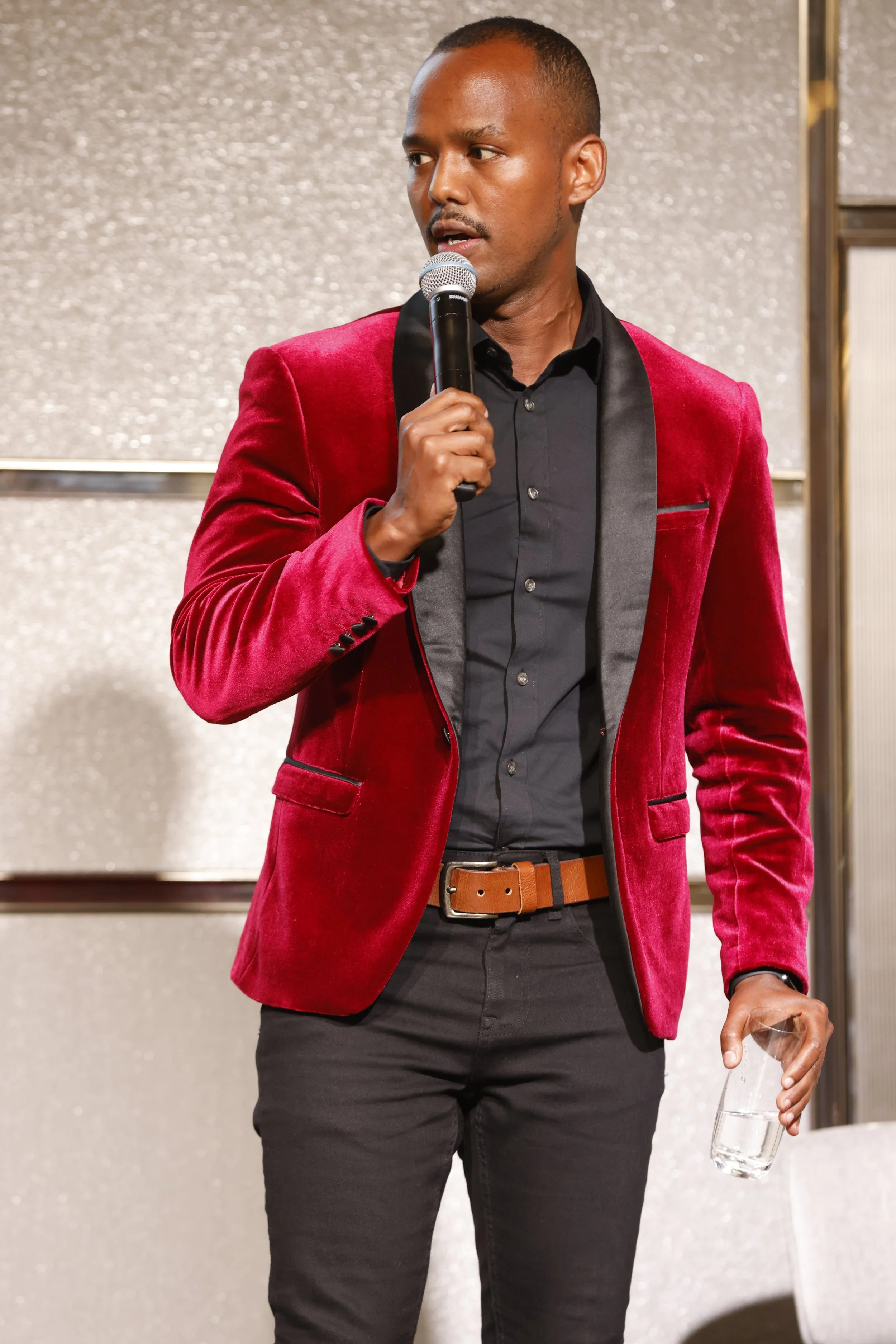 Man in red velvet blazer holding a microphone and glass of water, standing against a glittery wall.