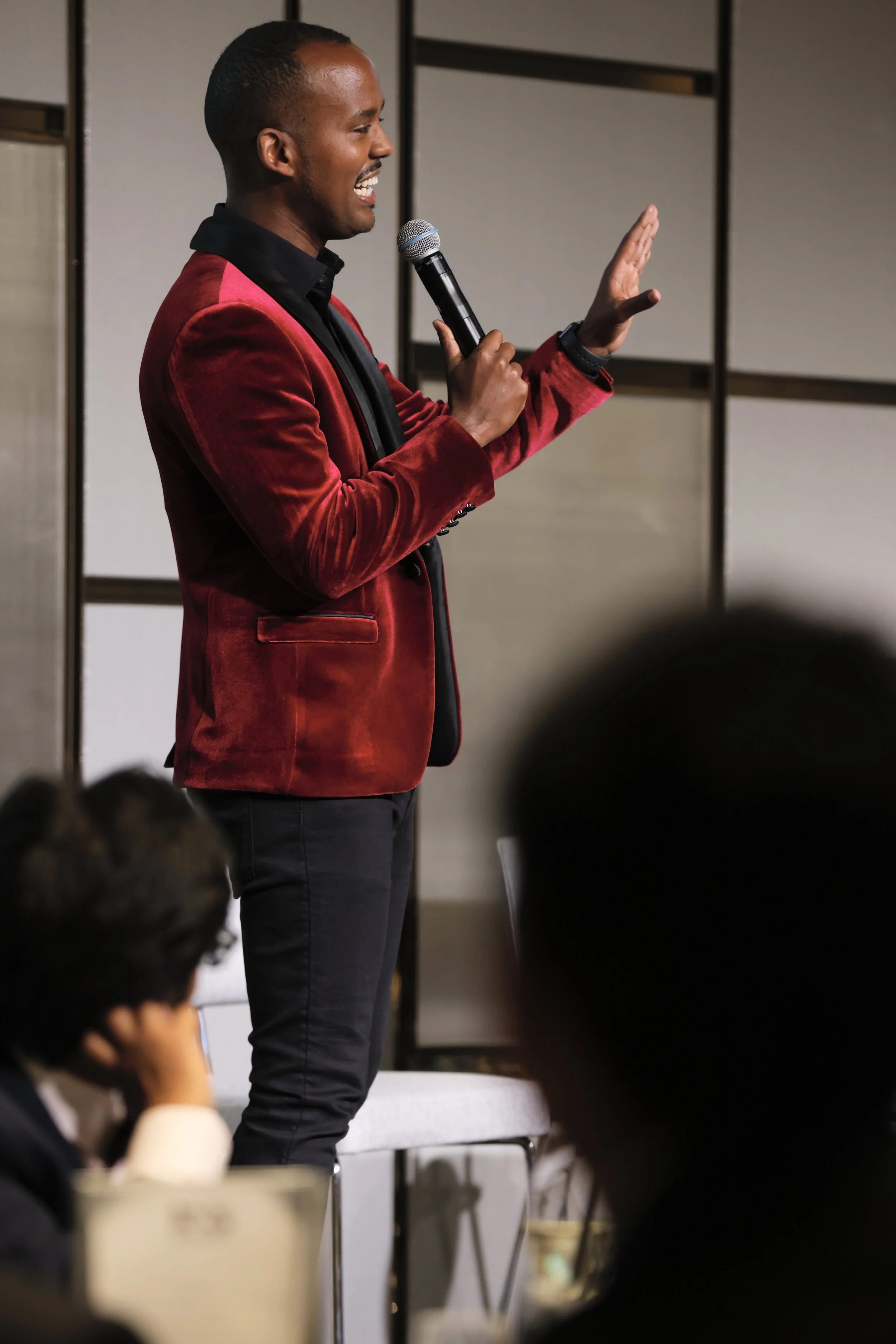 A man in a red jacket speaking or presenting with a microphone, smiling, in front of an audience.