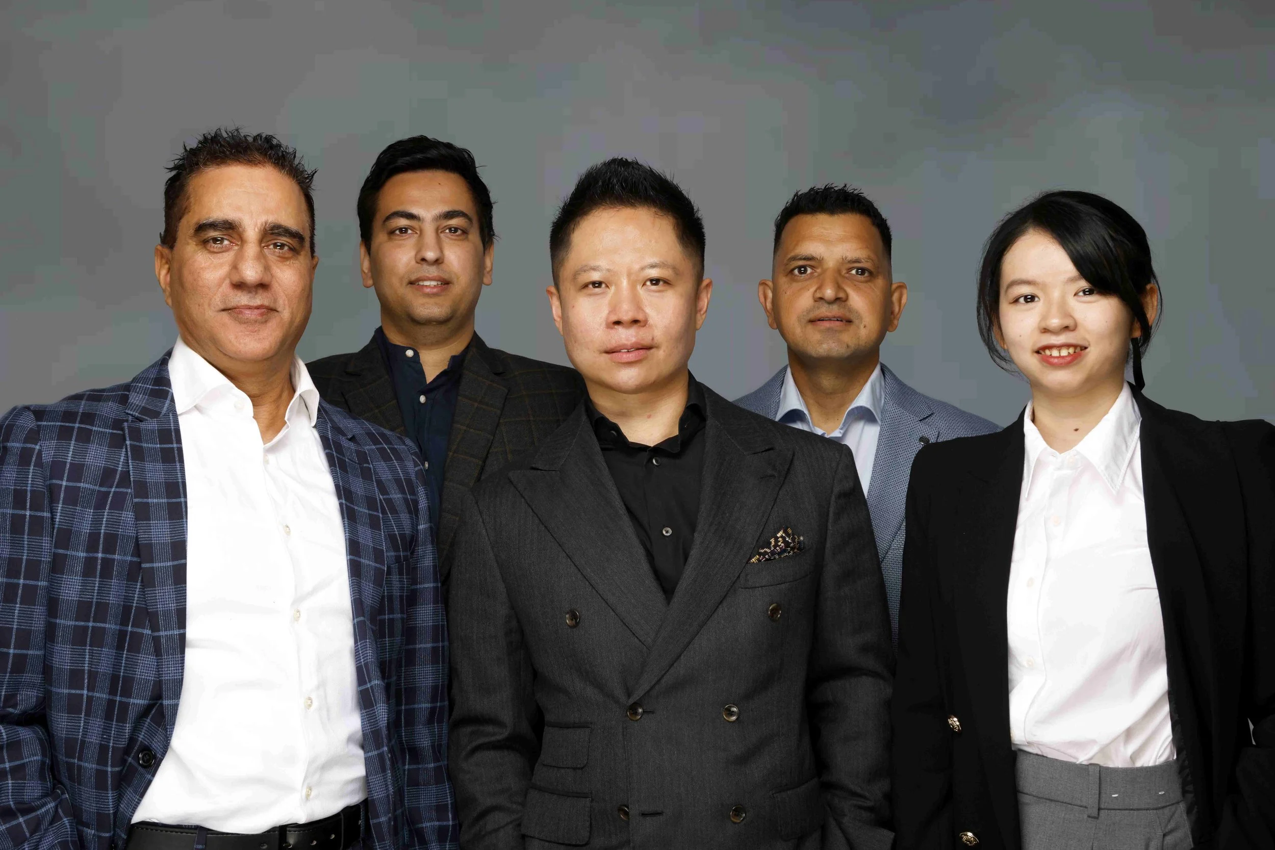 Group of five diverse business professionals in formal attire posing against a plain background.