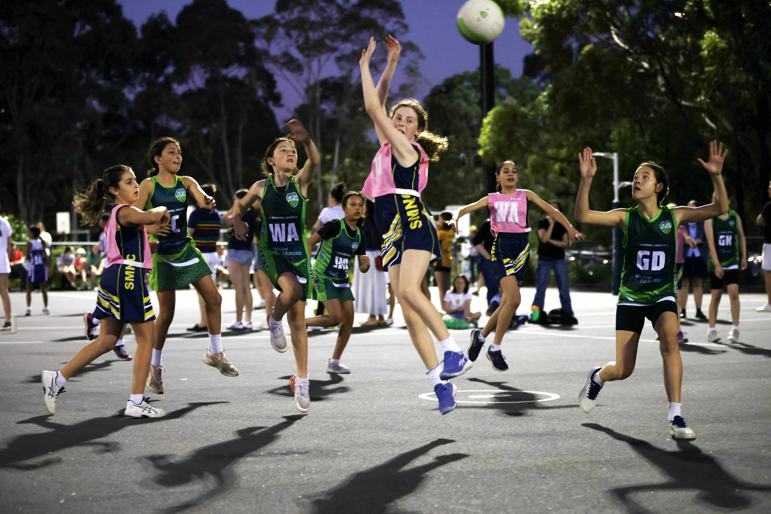 A group of young girls playing netball on an outdoor court. They are wearing green and pink uniforms, actively reaching for the ball. Trees and spectators are visible in the background.