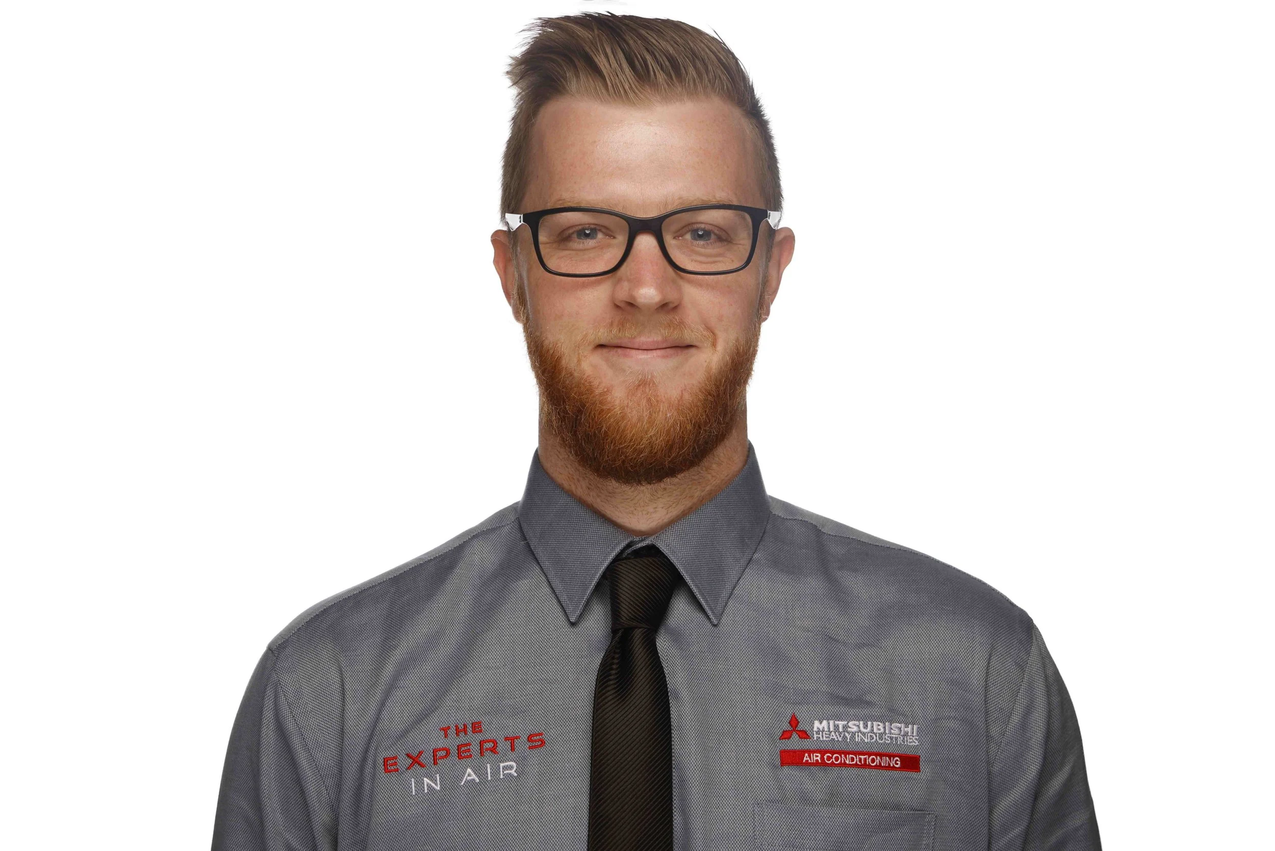 Portrait of a young man with glasses, red hair, and a beard, wearing a gray work shirt with company logos, against a white background.