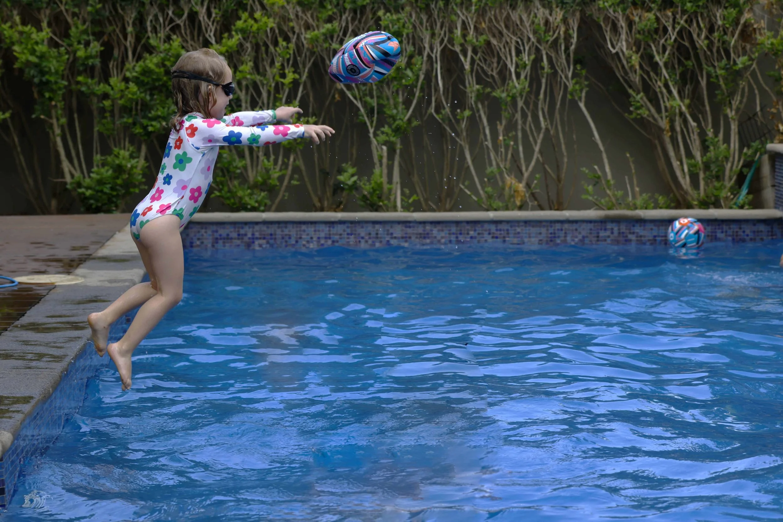 A young girl in a colorful floral bathing suit and sunglasses jumps into a swimming pool to catch a ball.