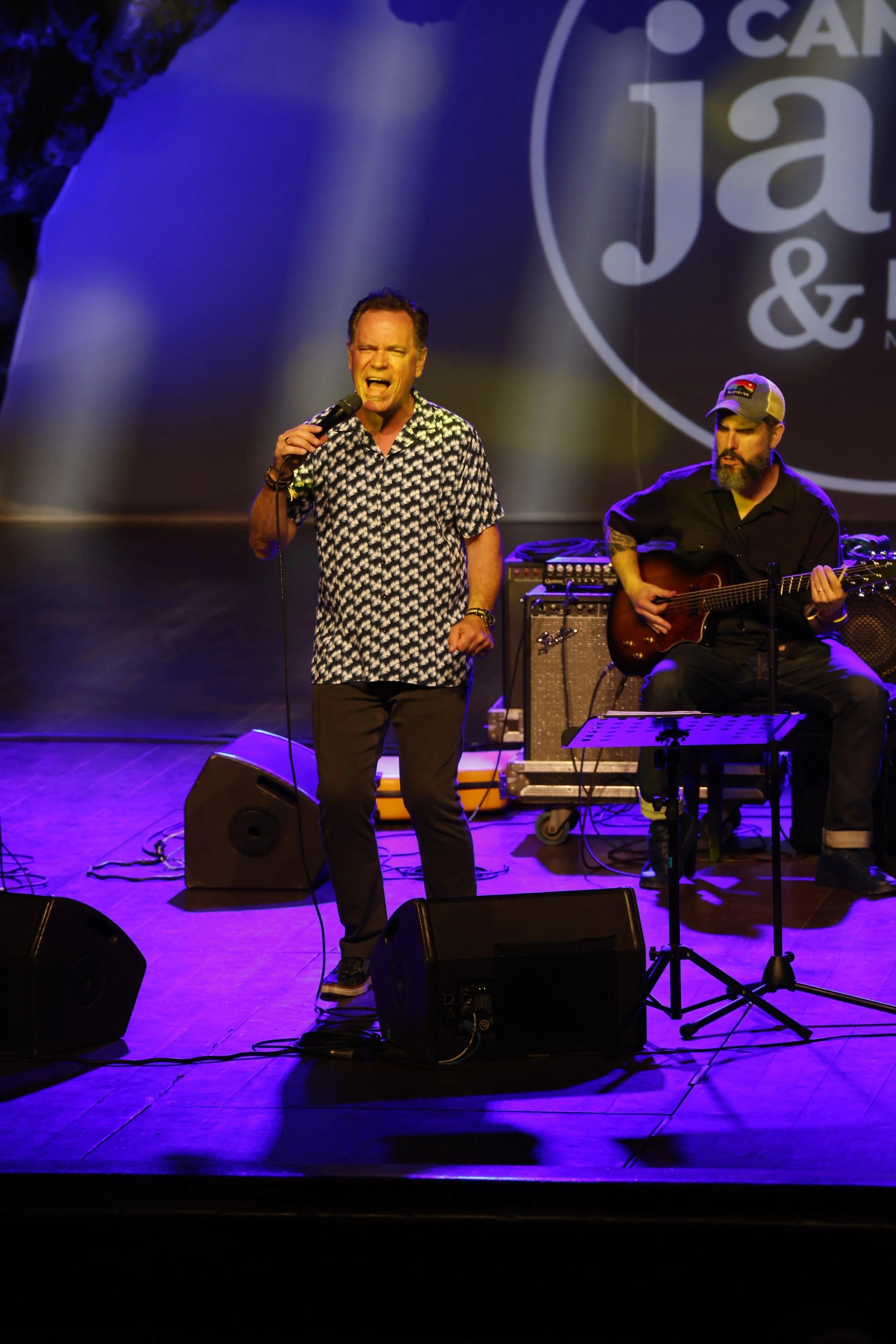 A man singing passionately into a microphone on stage, accompanied by another man playing an acoustic guitar, with stage lights and equipment visible, and a large logo in the background.