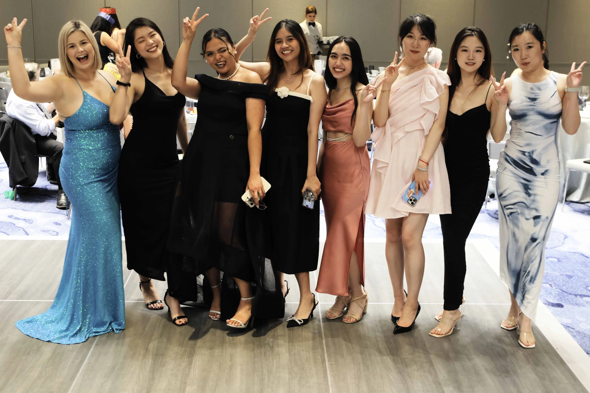 Group of nine women in formal dresses posing and smiling at an event.