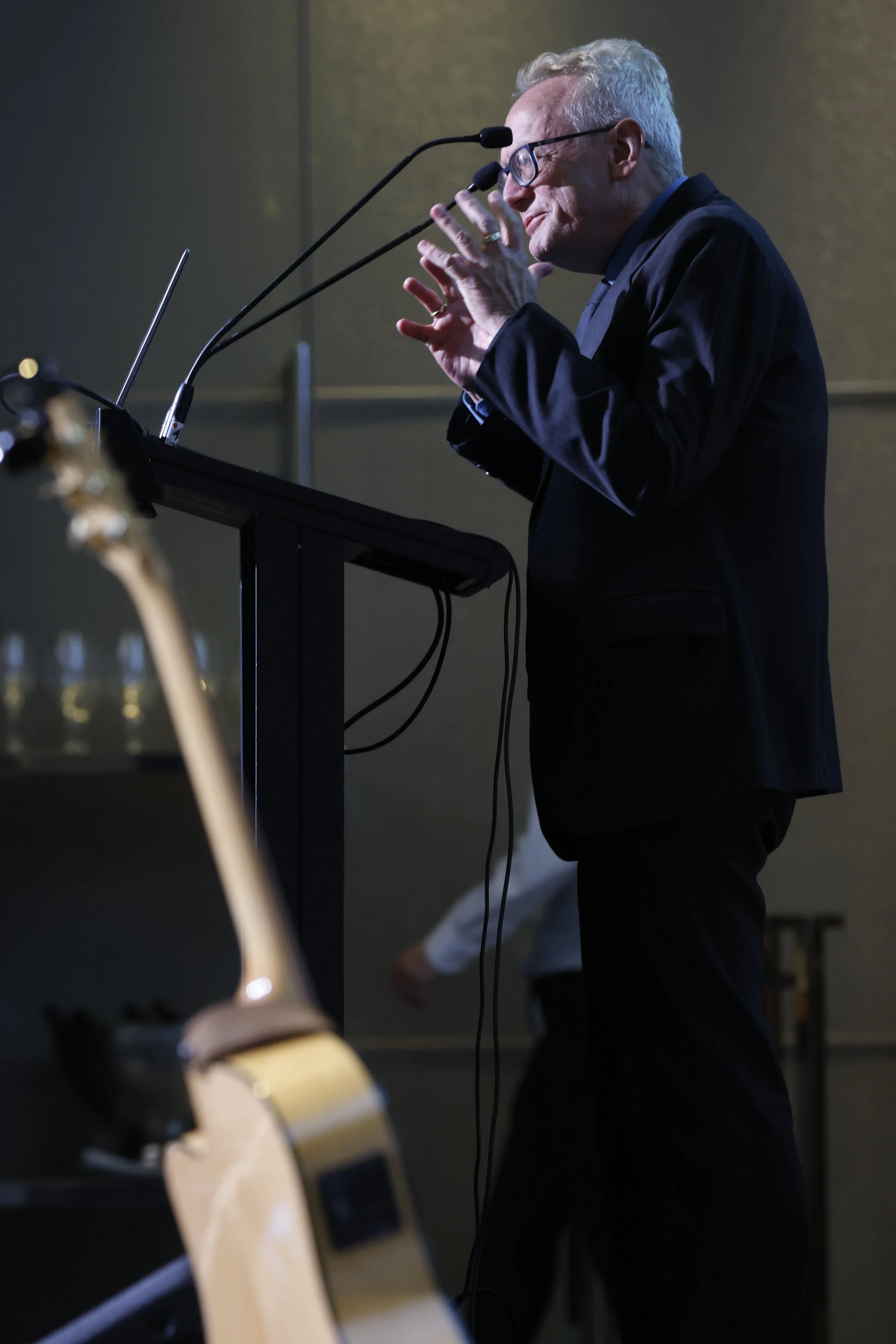 A man with glasses and white hair standing at a podium, speaking into two microphones. He is wearing a dark suit and is gesturing with his hands.