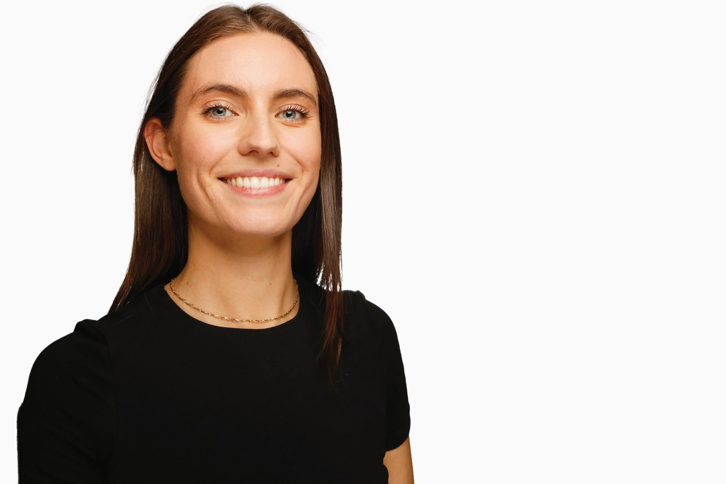 A woman with brown hair, blue eyes, and light skin, smiling, wearing a black shirt and a delicate gold necklace, against a plain white background.
