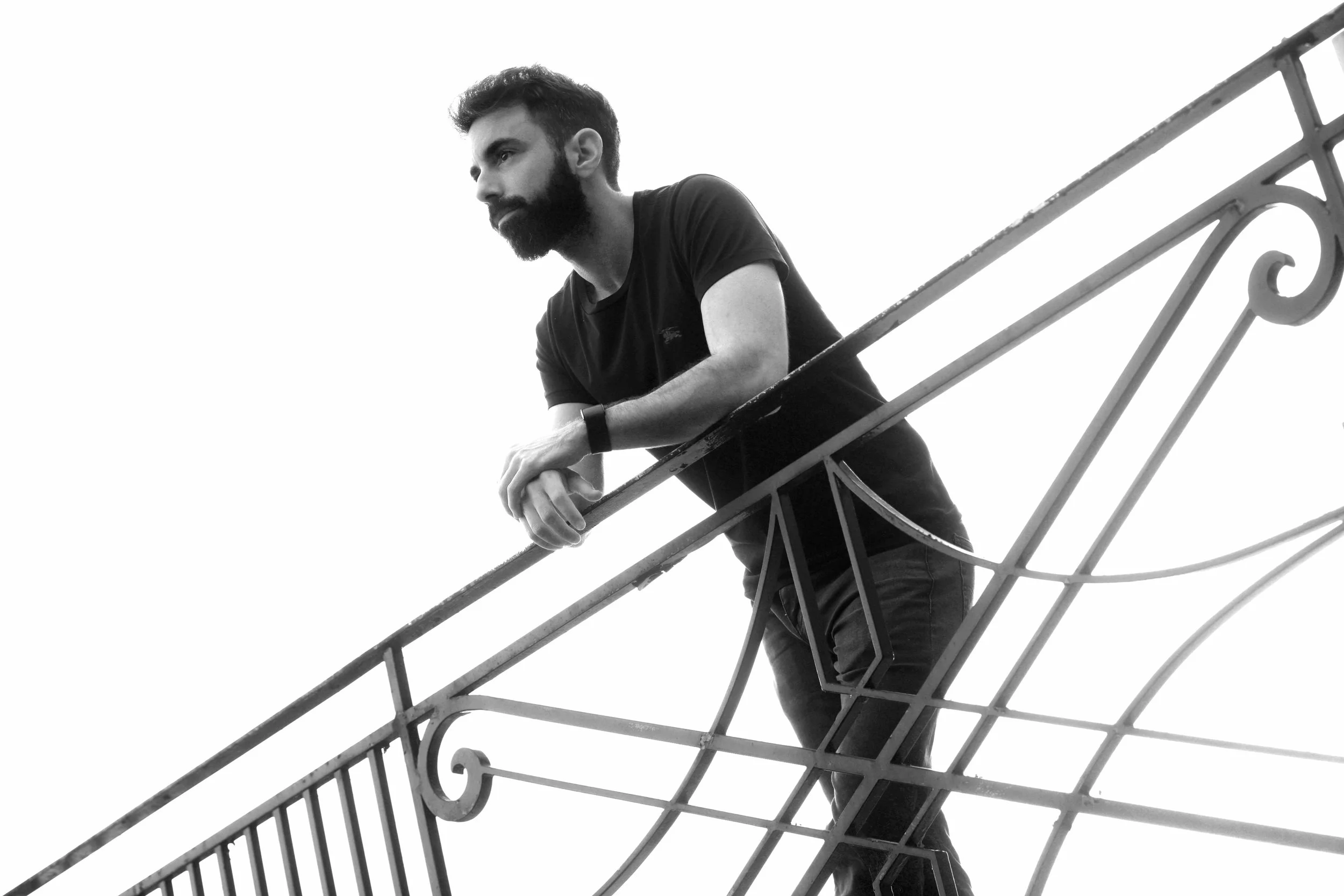 A man with a beard in a black t-shirt leaning on a decorative metal railing, looking thoughtfully into the distance, in black and white.