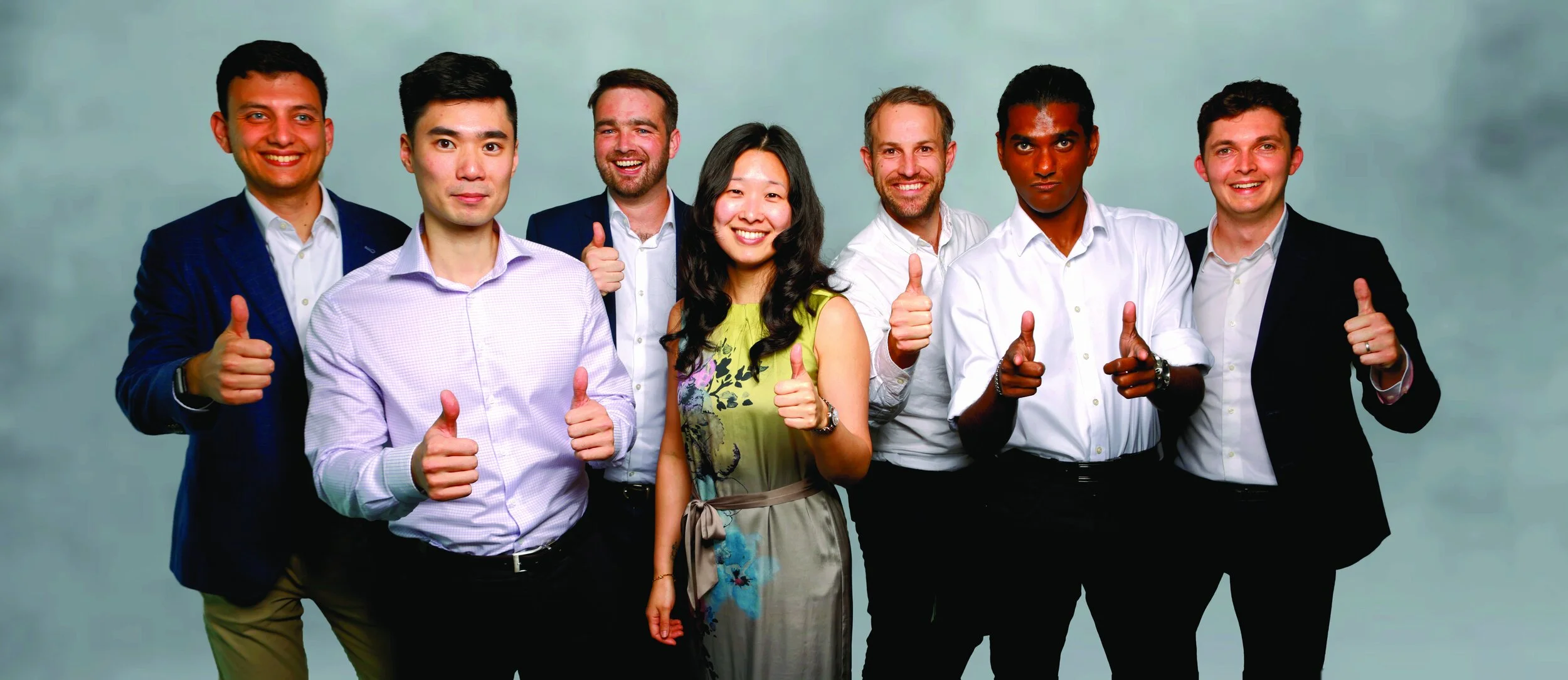Group of seven diverse professionals smiling and giving thumbs-up in a studio with a neutral background.