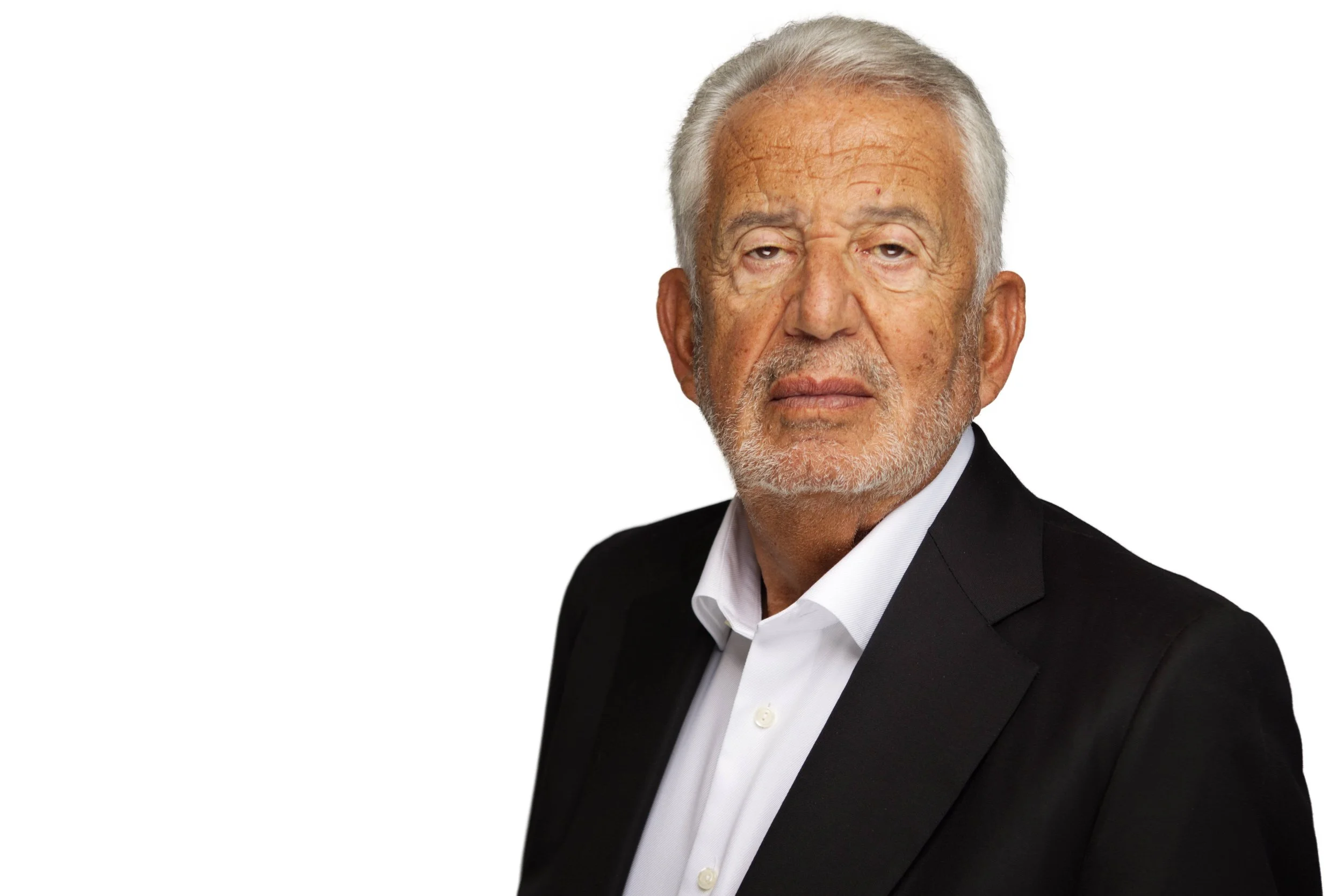 Portrait of an older man with gray hair and beard wearing a white collared shirt and black blazer, looking directly at the camera with a neutral expression.