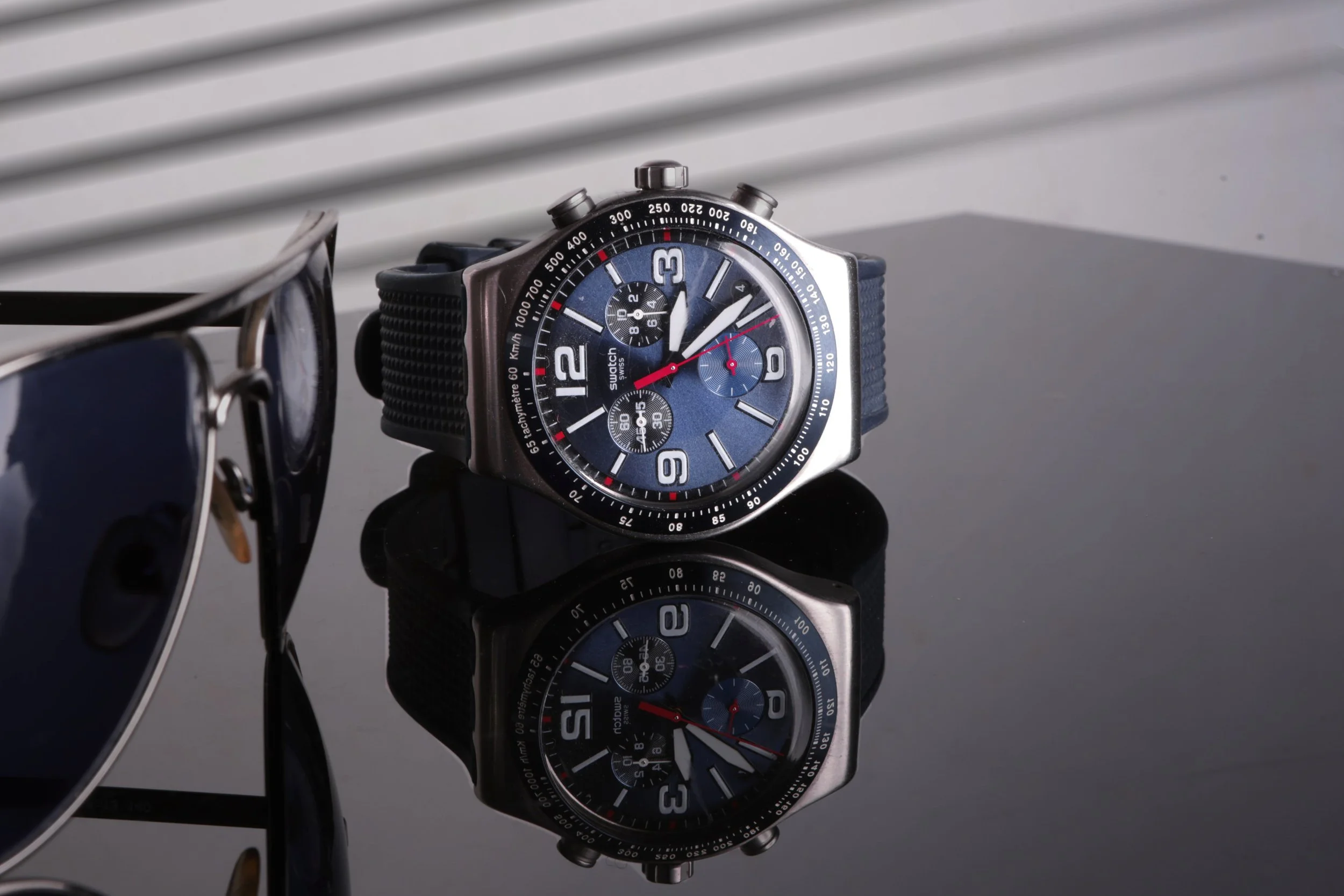 A wristwatch with a blue face and multiple dials, placed on a reflective dark surface, with a pair of sunglasses partially visible on the left.