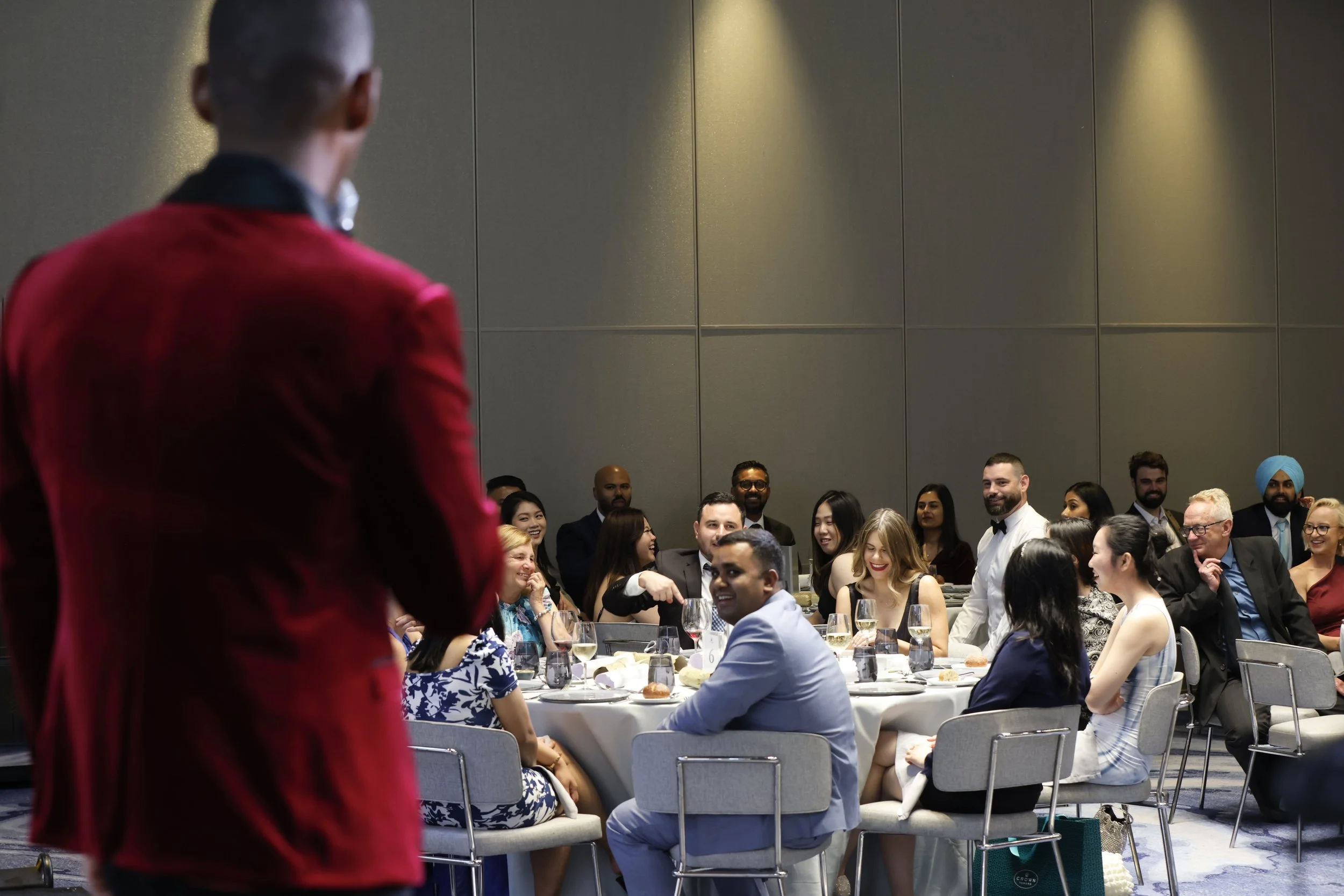 A man in a red suit speaking to an audience at a banquet with diverse guests seated around a table, smiling and engaging.