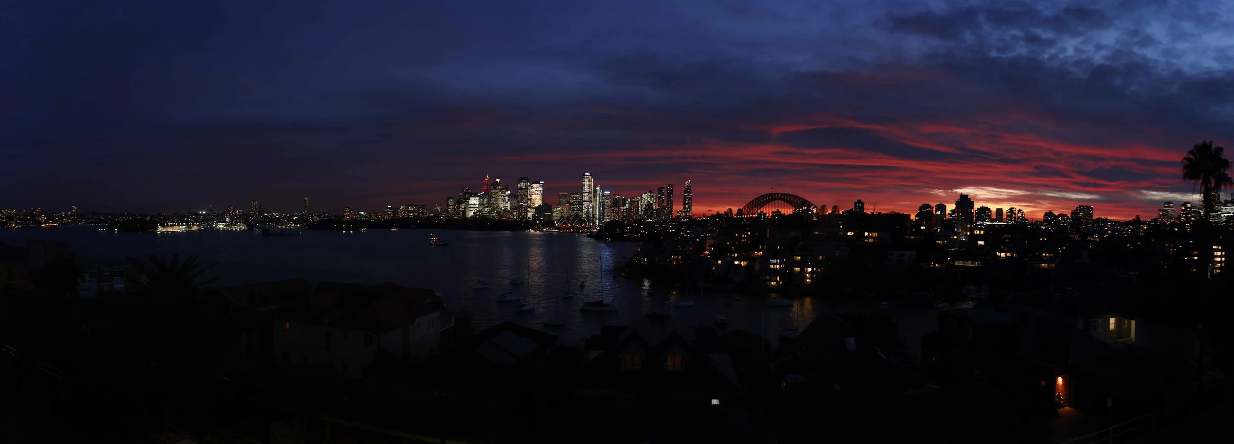 Nighttime city skyline view of a harbor with sailboats, illuminated buildings, and a prominent arch, under a colorful sunset sky.