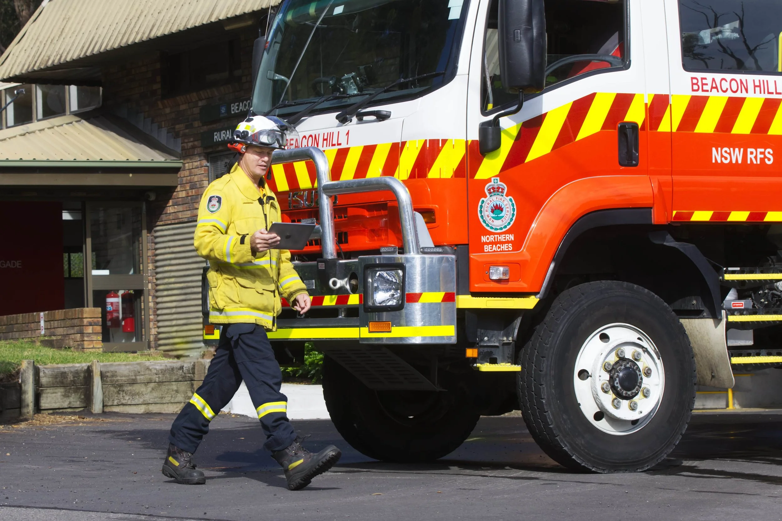 A firefighter in yellow uniform walking past a large red fire truck with yellow and red stripes, labeled 'Beacon Hill 1' and 'NSW RFS', parked on the street in front of a building.