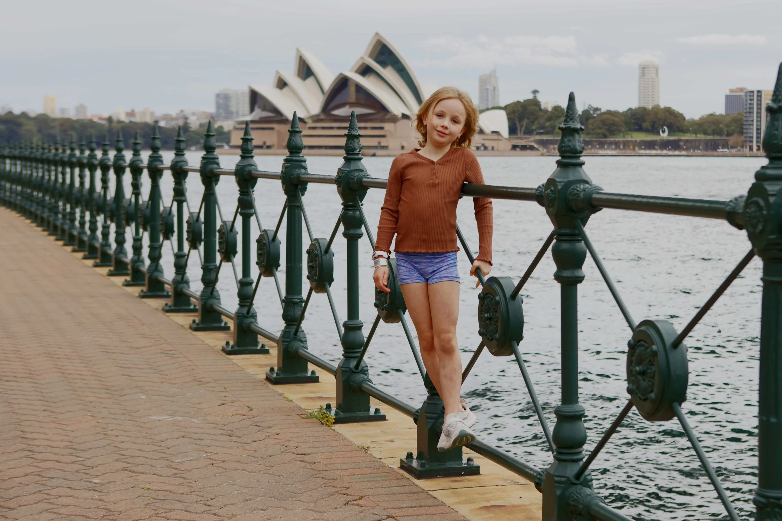 A young girl with red hair in a brown long-sleeve shirt and purple shorts, walking along a waterfront promenade, with the Sydney Opera House visible in the background.