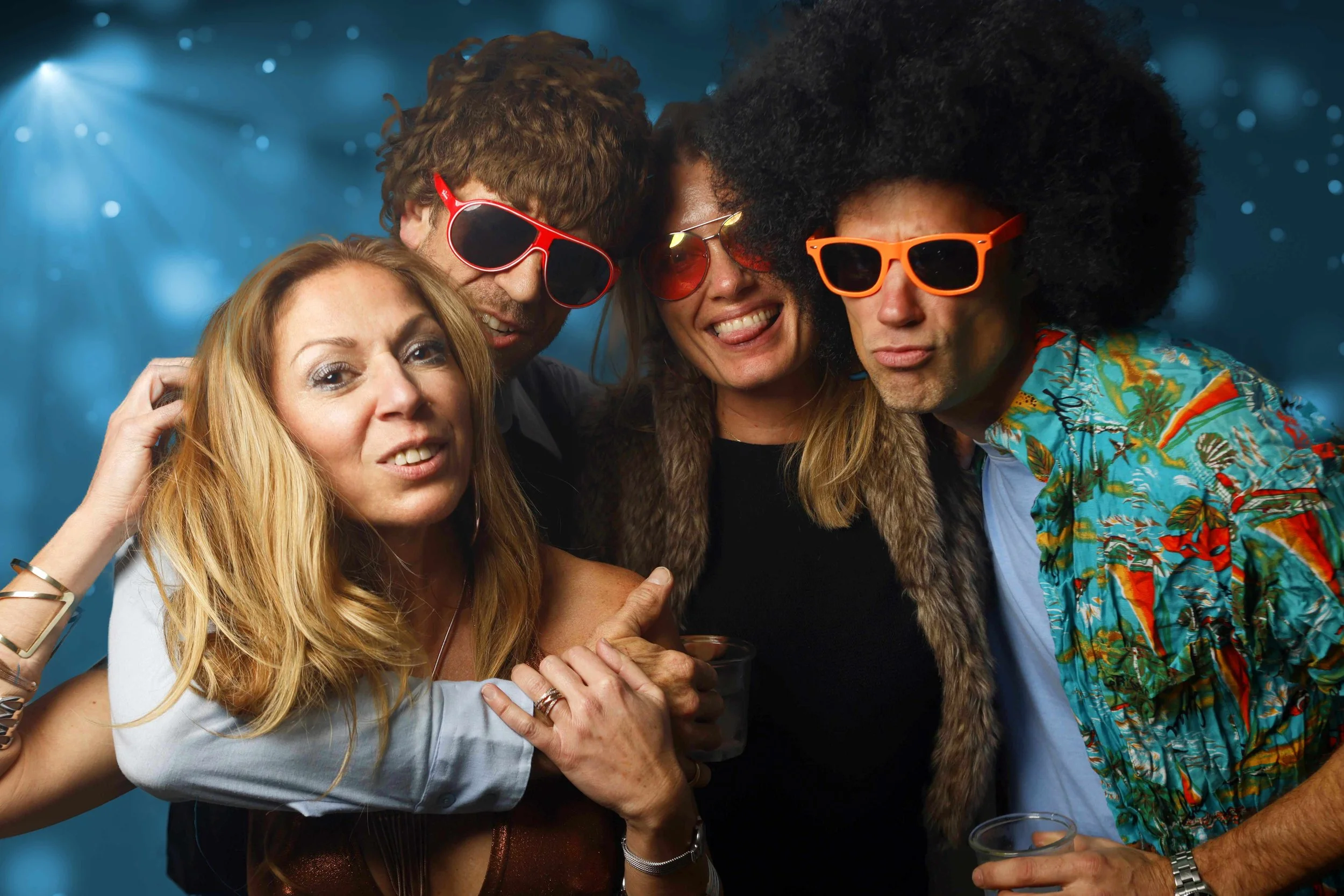 Group of four friends at a party wearing colorful sunglasses, smiling, and posing for the camera against a blue, bokeh background.
