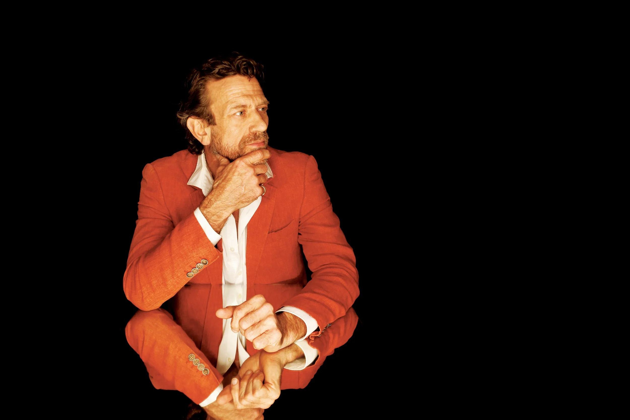 Thoughtful middle-aged man with beard wearing an orange suit and white shirt, sitting with his hand on his chin against a black background.
