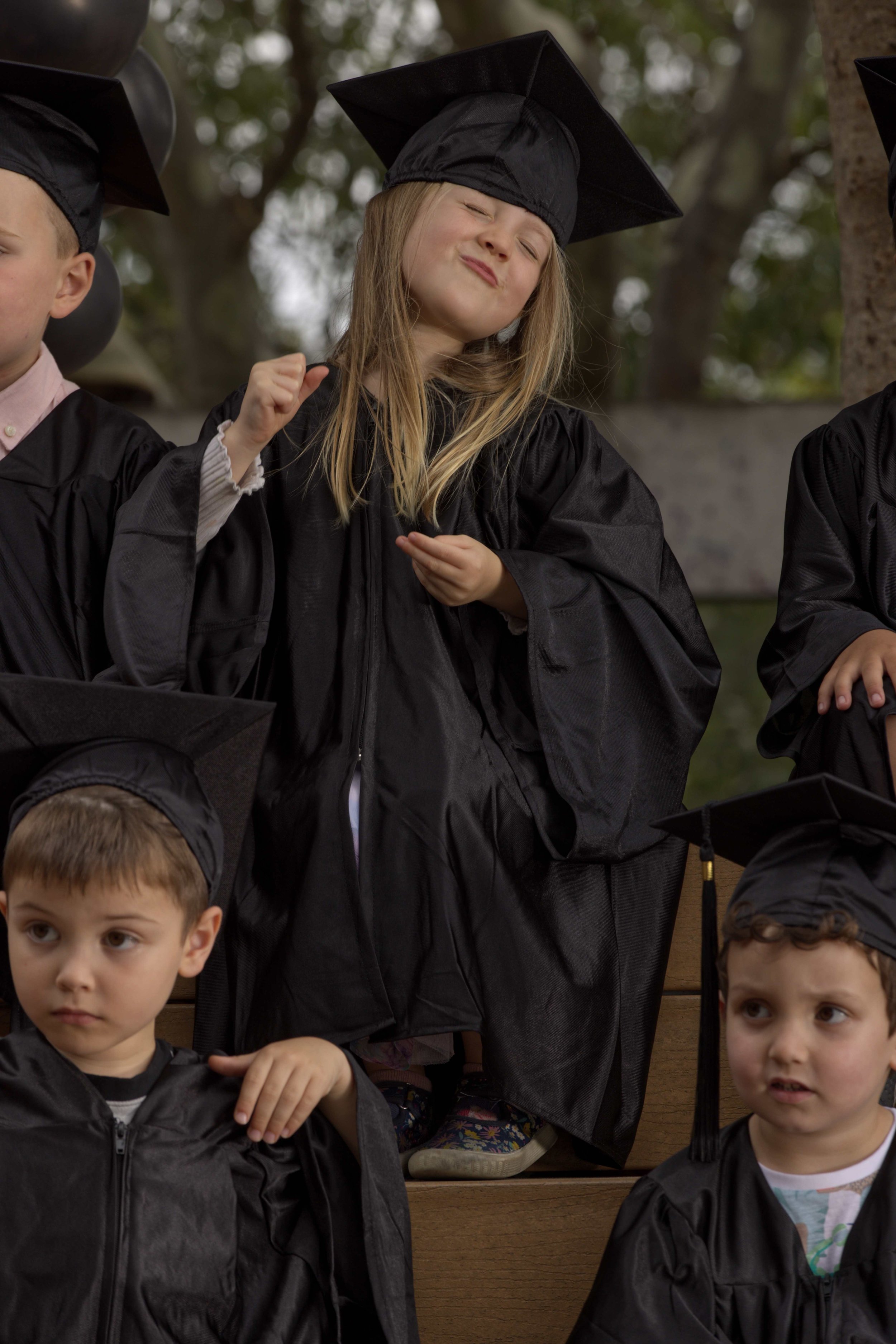 Three young children wearing black graduation gowns and caps, sitting on wooden steps, with one child in the center making a playful expression.