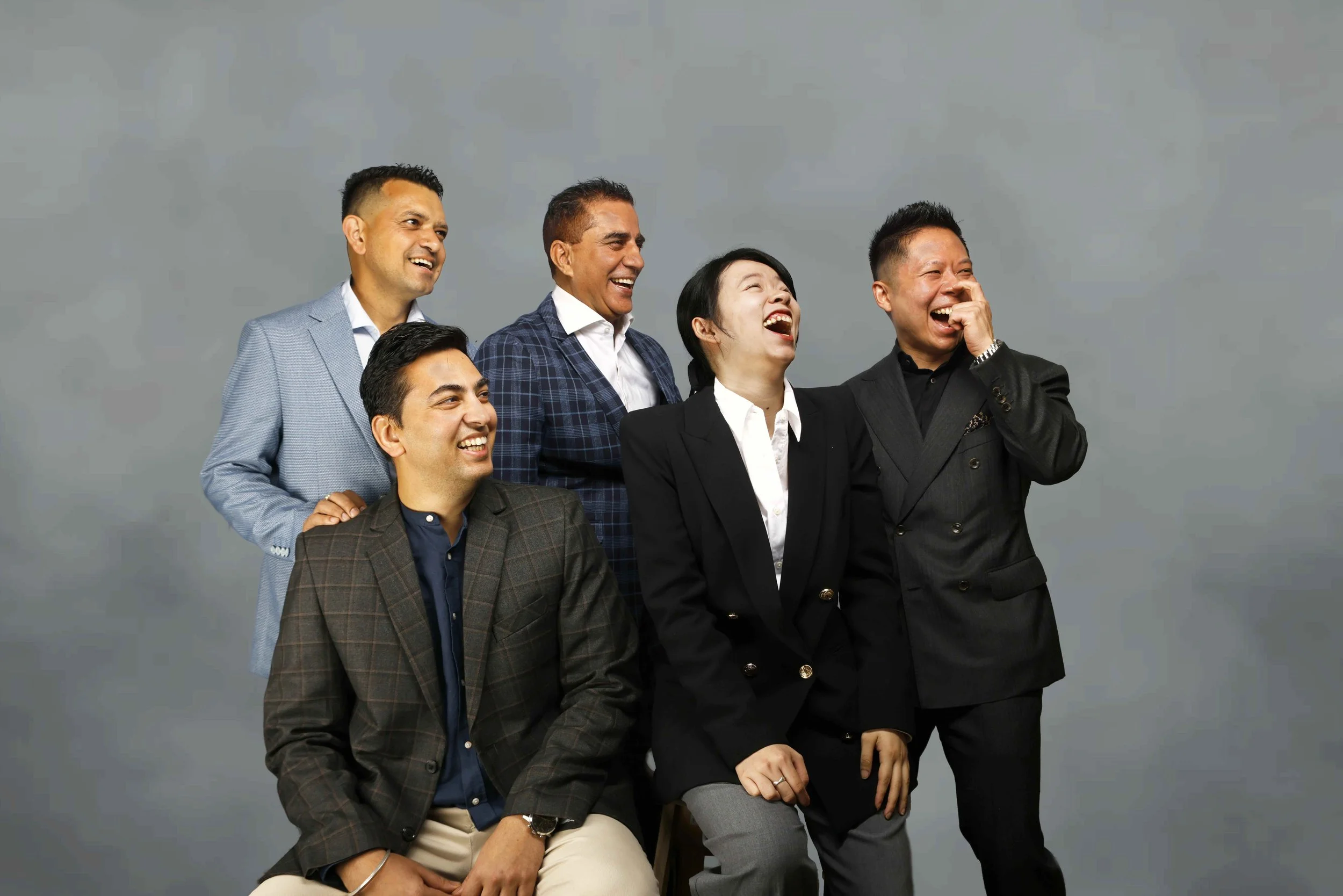 Group of six diverse professionally dressed people laughing and smiling against a gray background.