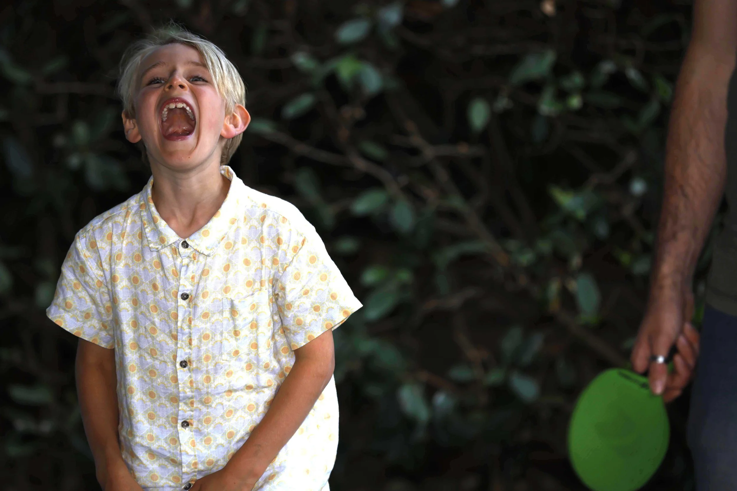 A young boy with blonde hair, wearing a yellow patterned shirt, is laughing with his mouth wide open. To the right, part of an adult is visible, holding a green paddle.