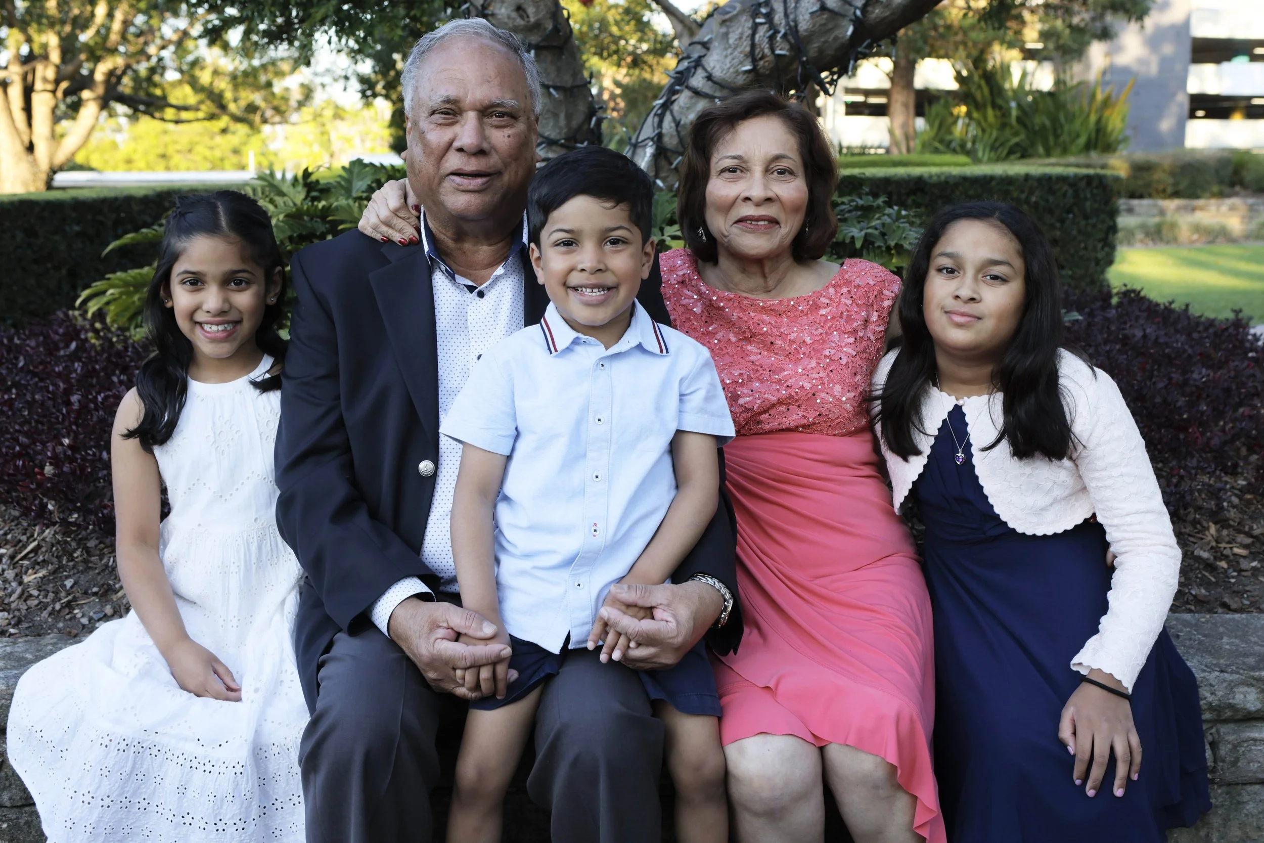 Family of six sitting together outdoors in a garden, including grandparents, two young girls, and a young boy, all smiling.