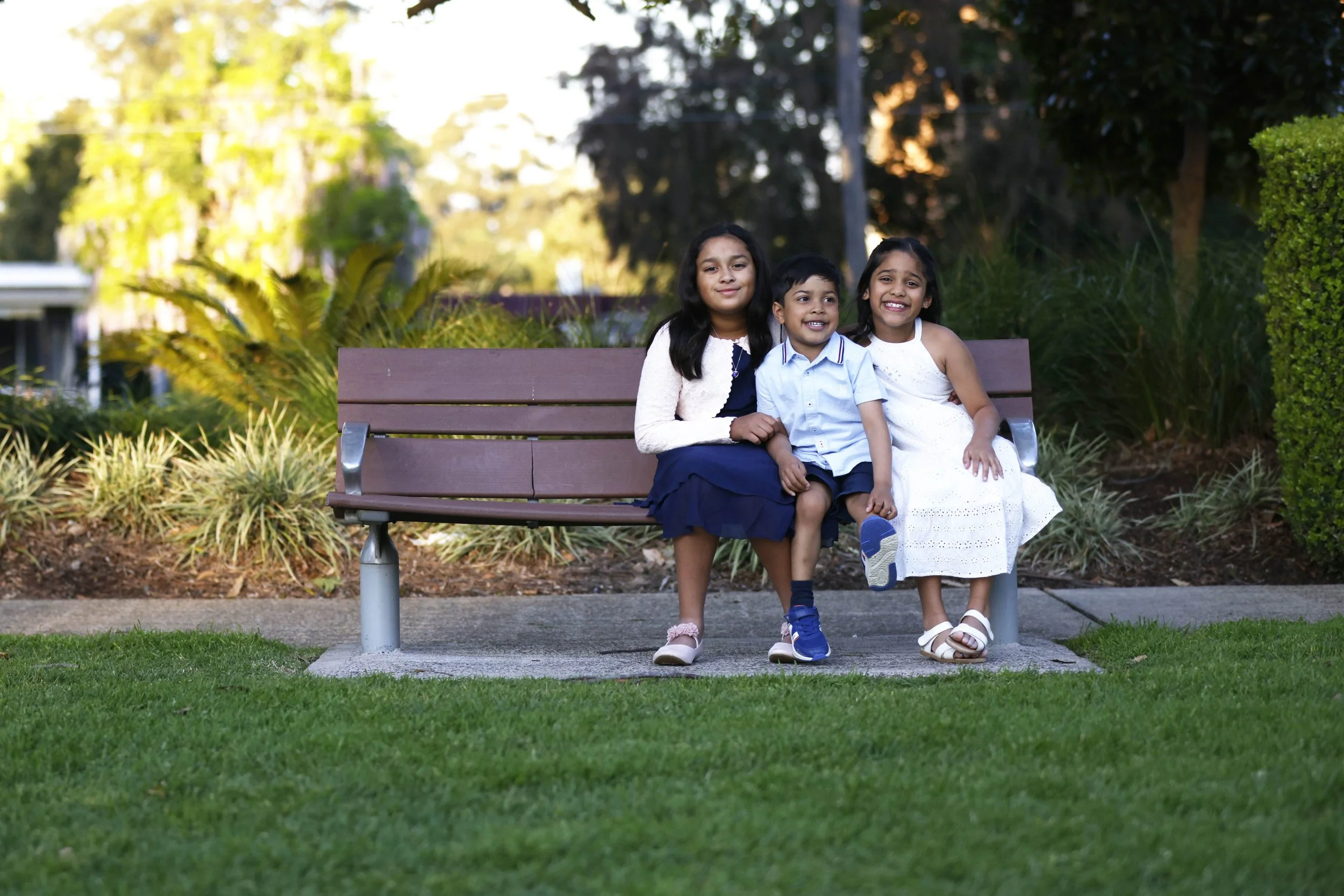Three children sitting on a park bench, smiling, with greenery and trees in the background.