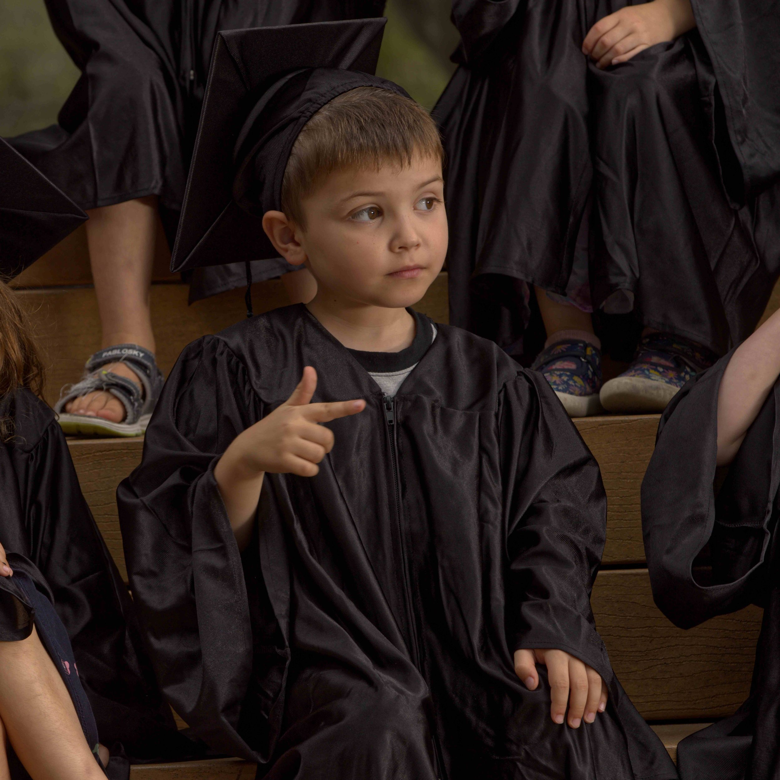A young child in a black graduation gown and cap sitting on a wooden bench, making a gesture with their fingers. Other children in similar attire are partially visible in the background.
