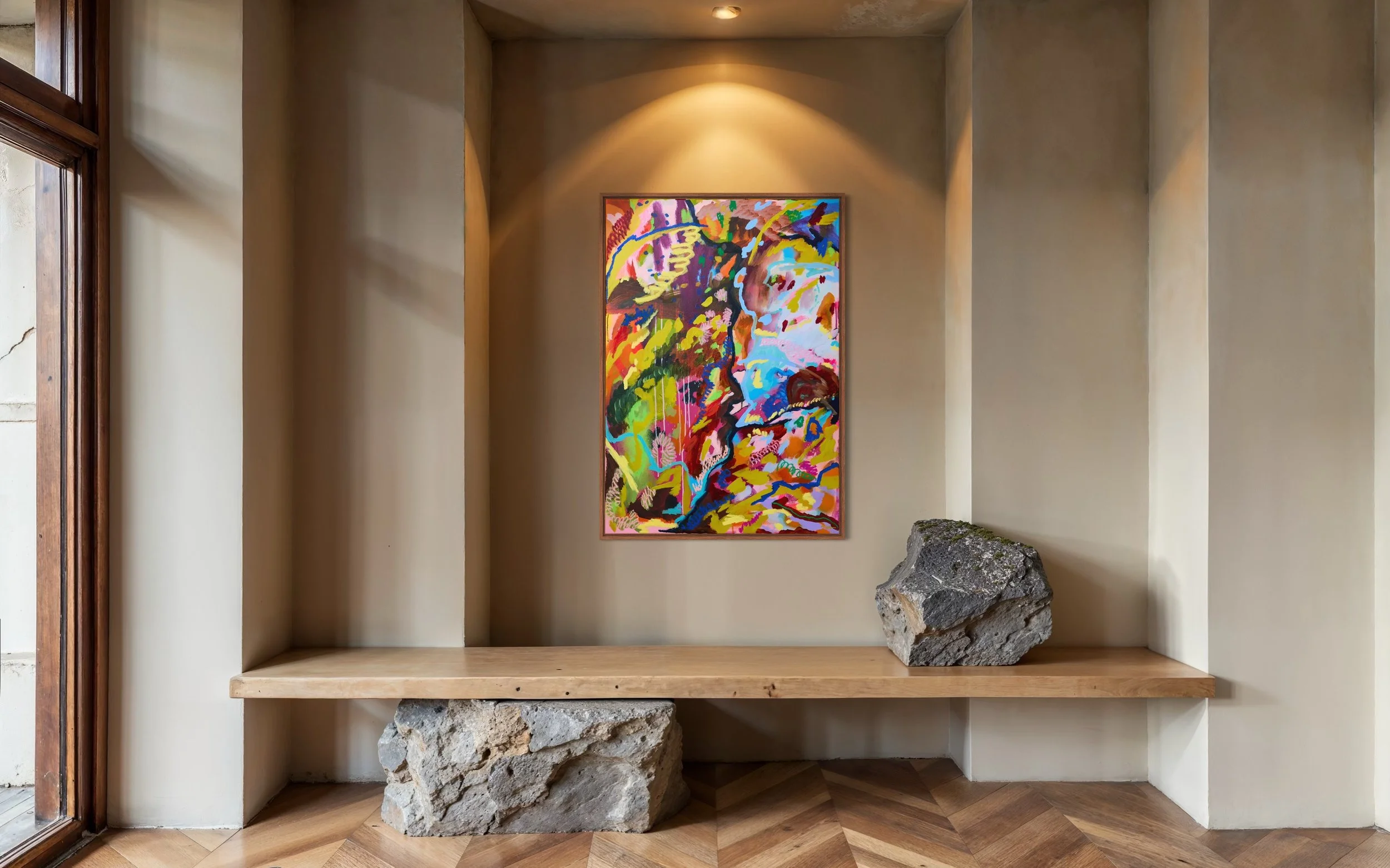 A colorful abstract painting hangs on a beige wall inside a room, illuminated by overhead lighting. A wooden shelf below the painting holds two large stones, one on each end. A window with wooden trim is on the left side of the image.