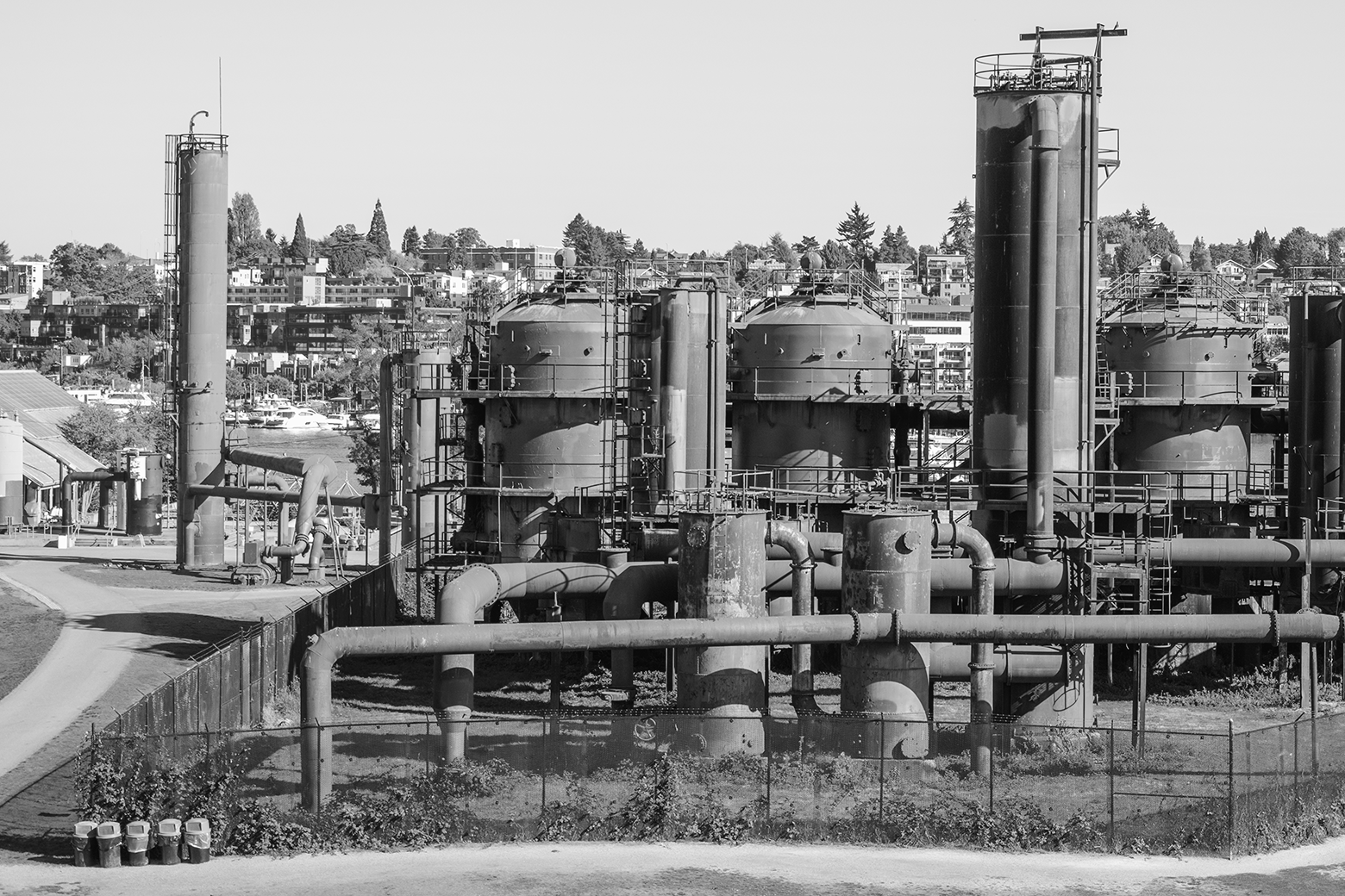 Black and white photo of an industrial site with large storage tanks, pipes, and walkways.