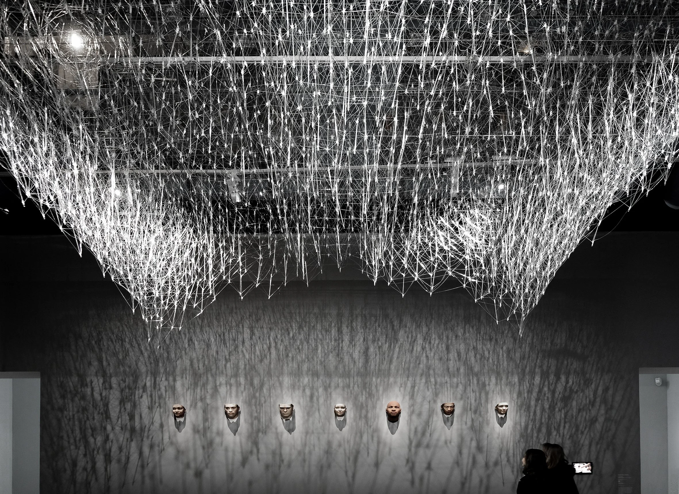 An art installation with a large, intricate, web-like hanging structure illuminated from above, casting shadow patterns on the wall below.