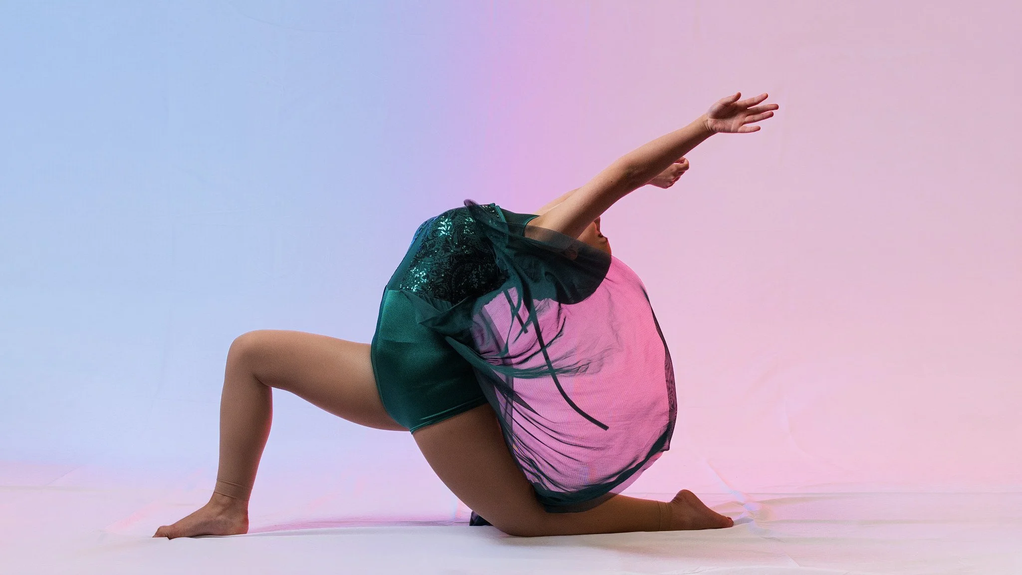 A person in a pose combining yoga and dance elements, dressed in a shiny, black and green top and shorts, on a light background with a gradient of pink and blue. Thetford dance photography by butterflies k-photography.