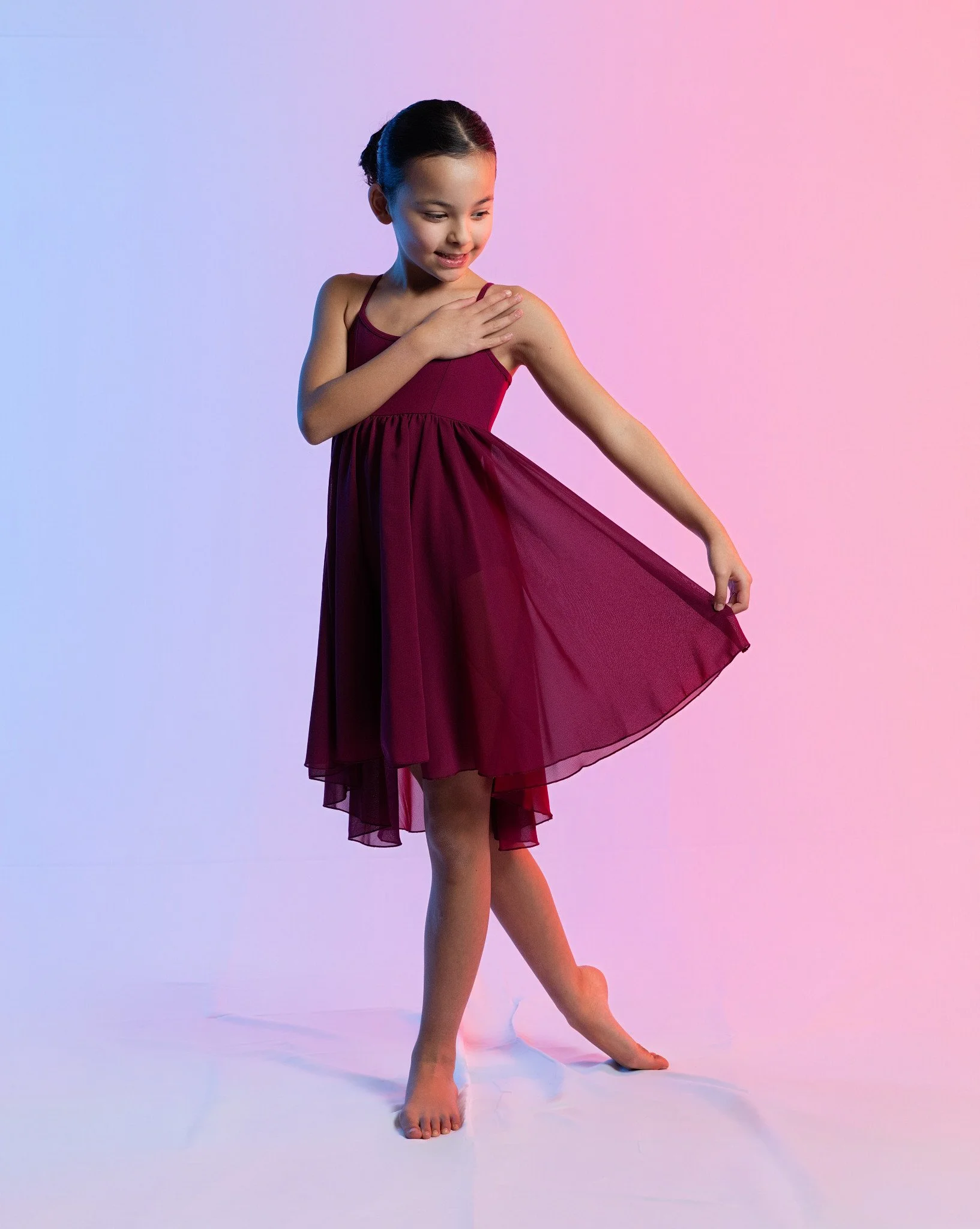 A young girl in a burgundy dress standing on a colorful pink and blue gradient background, smiling and looking down at her dress while holding it out with one hand. Thetford dance photography by butterflies k-photography.