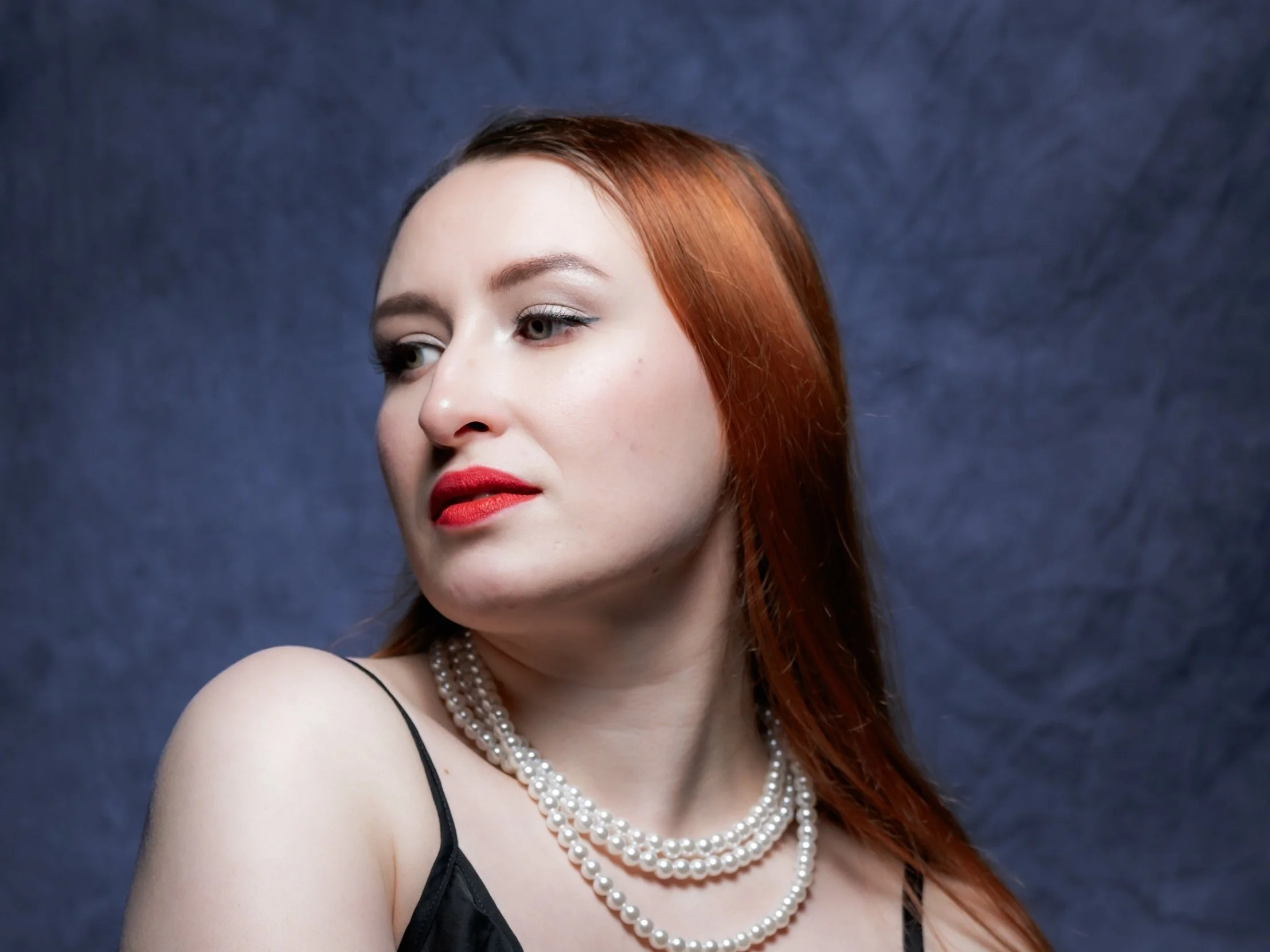 A woman with red hair and fair skin wearing red lipstick, pearl necklaces, and a black dress, looking to her left against a dark textured background. Portrait by butterflies k-photography Thetford.