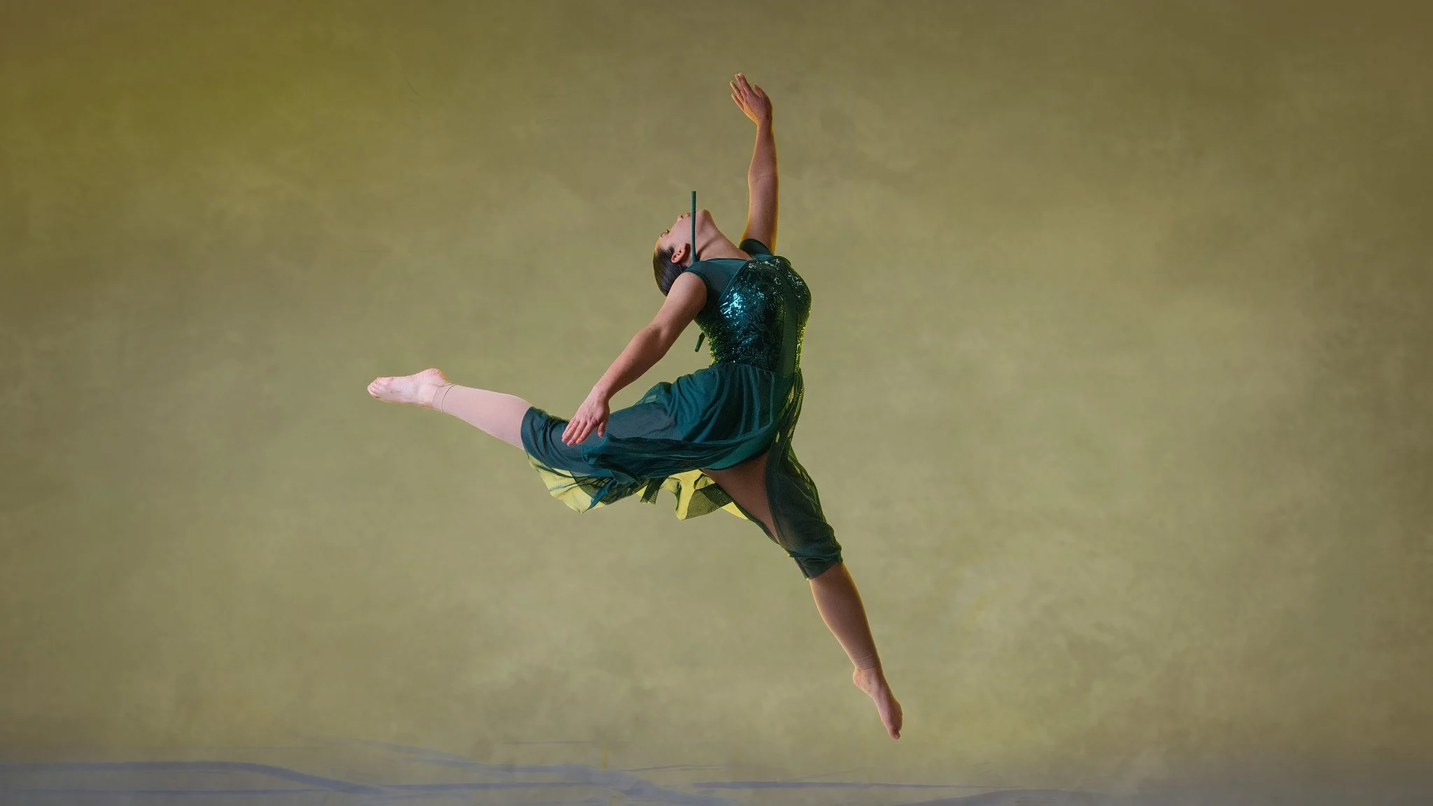 A female ballet dancer is mid-air, performing a leap with one leg extended forward and the other backward, wearing a dark green dress with a sequined top and a sheer skirt. Thetford dance photography by butterflies k-photography.