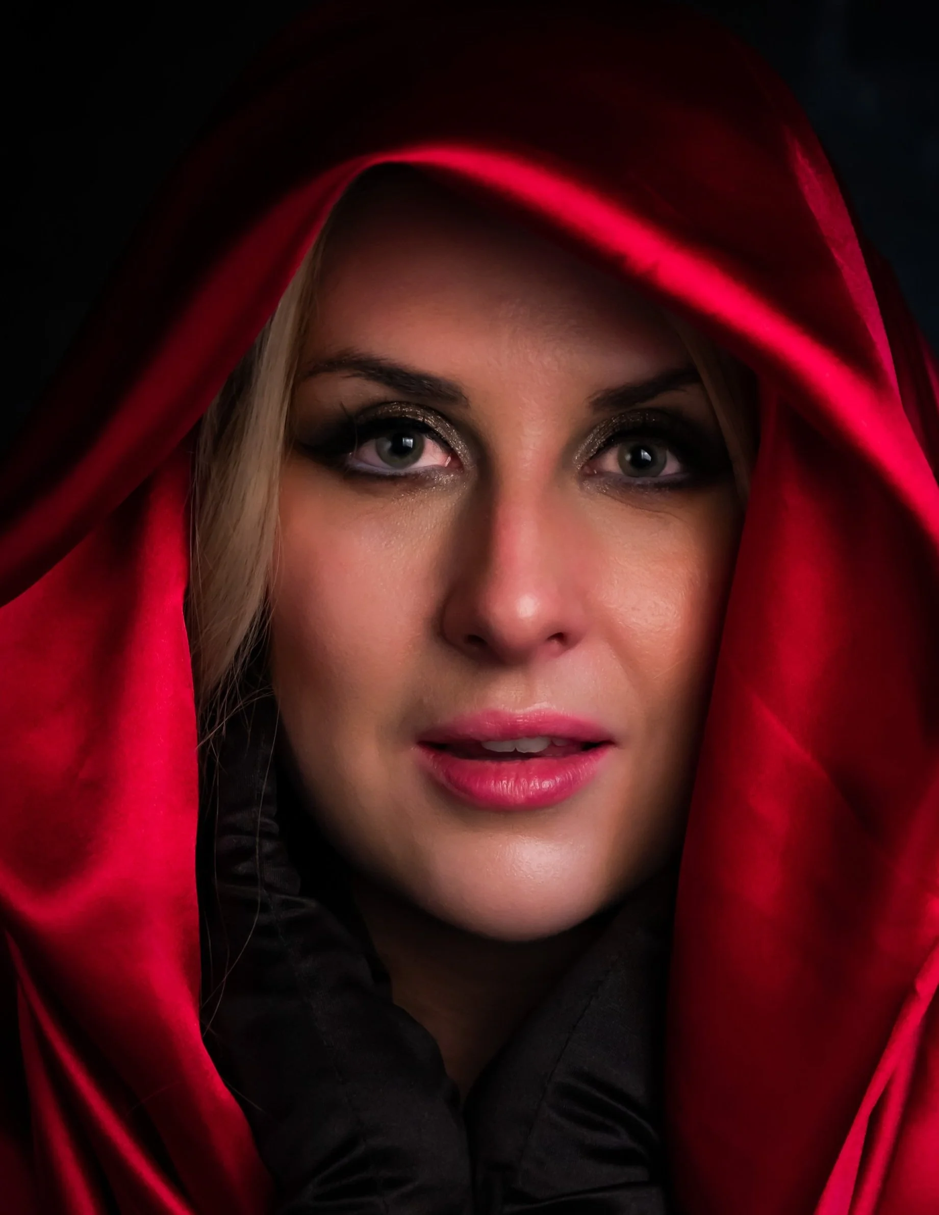 Close-up of a woman with light skin wearing a red hooded cloak, dark eye makeup, and pink lipstick, looking directly at the camera. Portrait by butterflies k-photography Thetford.