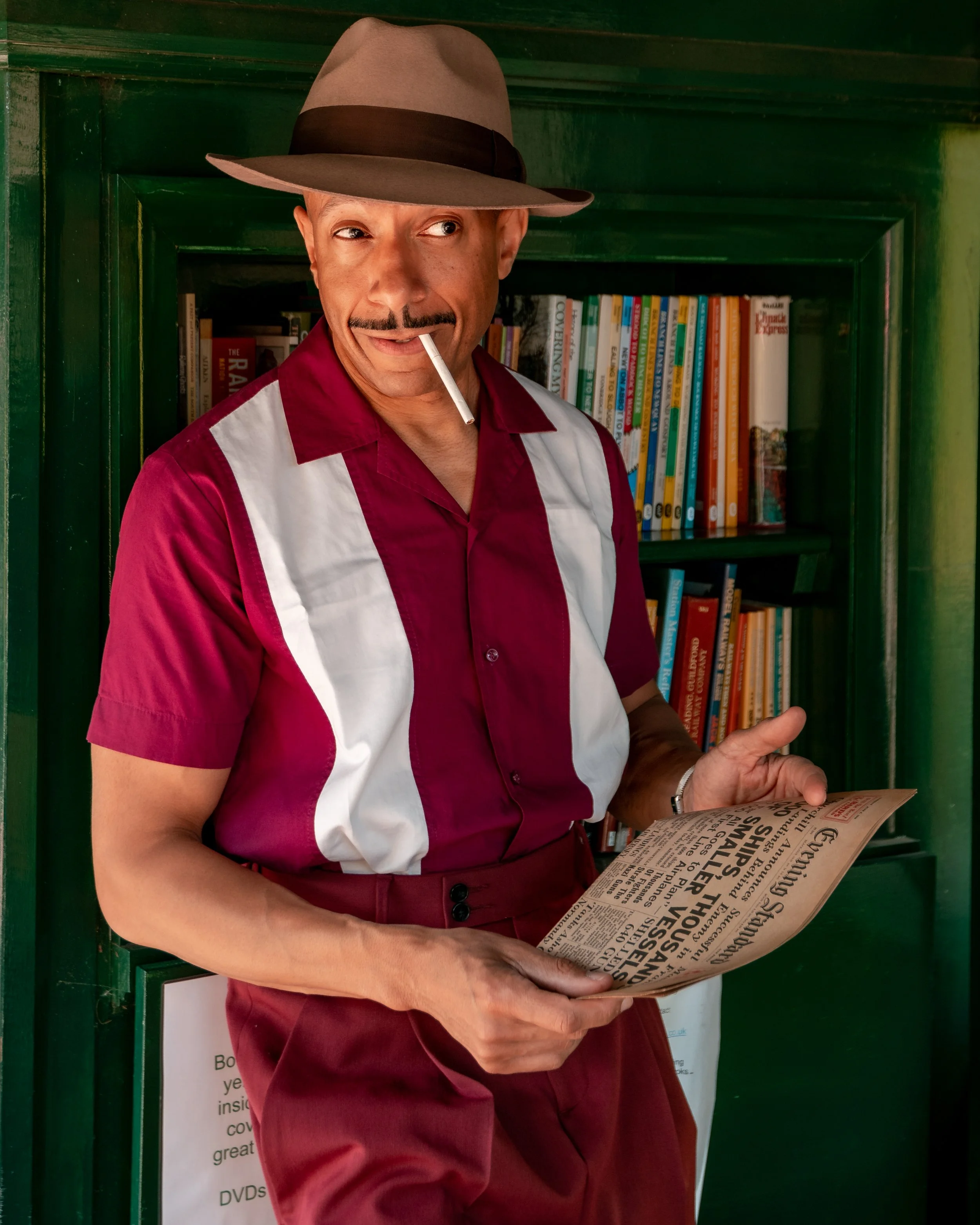 A man wearing a brown fedora, red and white short-sleeved shirt, and red pants, holding a newspaper, with a cigarette in his mouth, standing in front of a green bookshelf filled with books. Portrait by butterflies k-photography Thetford.