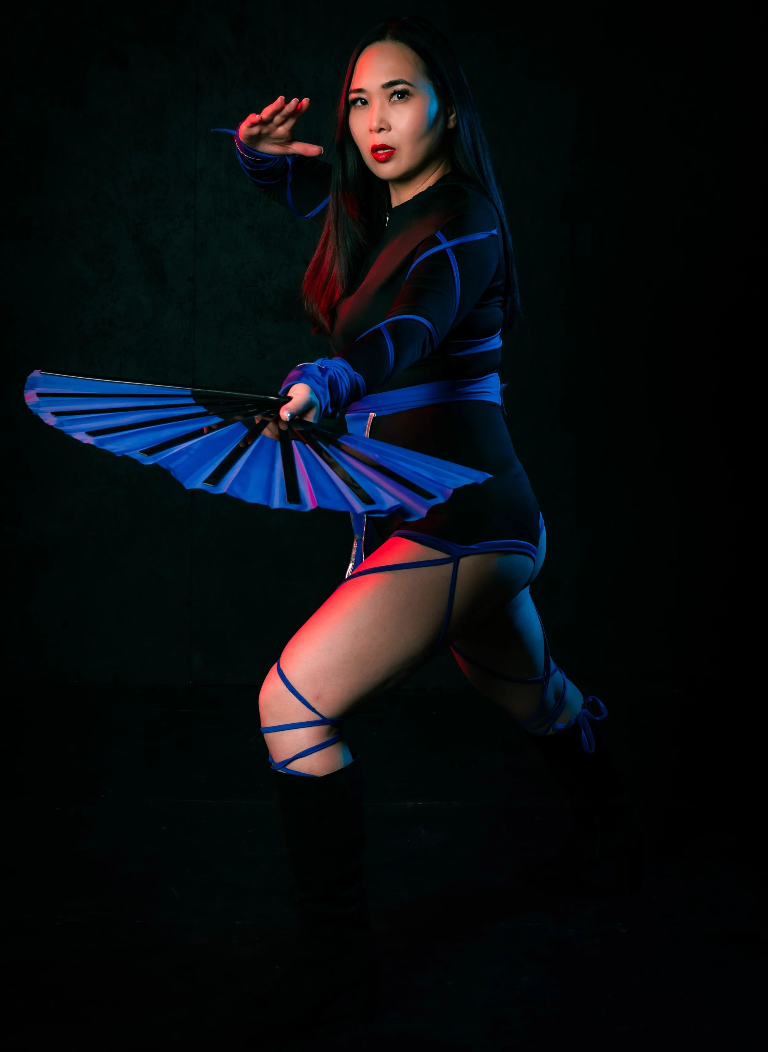 A woman dressed in a black and blue costume holding a fan, striking a pose against a dark background with red and blue lighting. Portrait by butterflies k-photography Thetford.