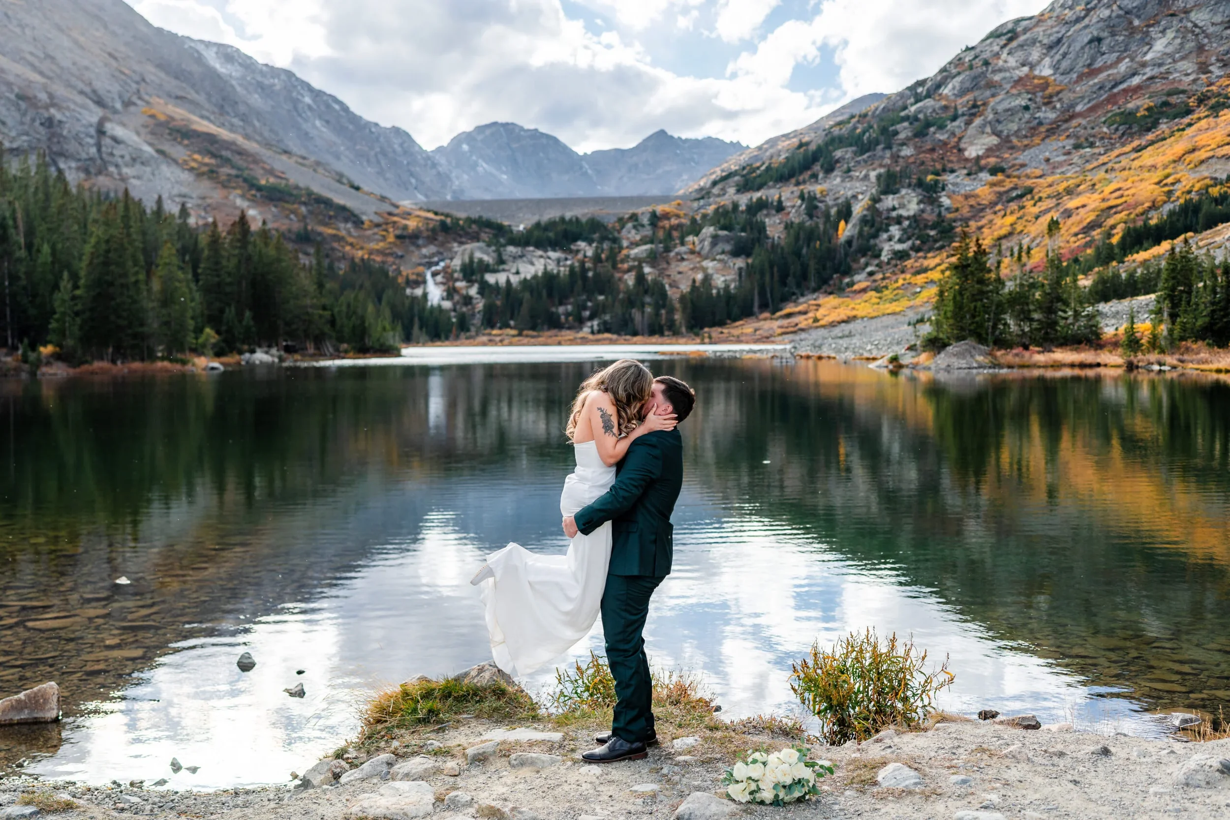 A couple in wedding attire sharing a kiss and embracing in front of a mountain lake with trees and mountains in the background, during daytime.