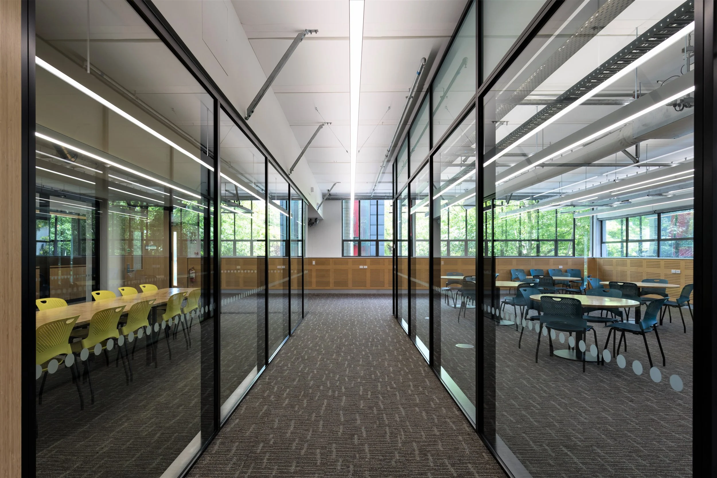 View of a modern conference or meeting room with glass walls, tables, and chairs, with large windows showing trees outside.