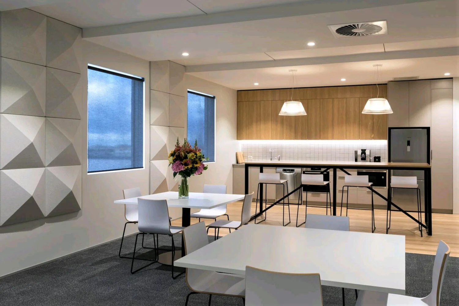 Modern kitchen and dining area with white tables, black chairs, a vase of colorful flowers, wooden cabinets, and white pendant lights.