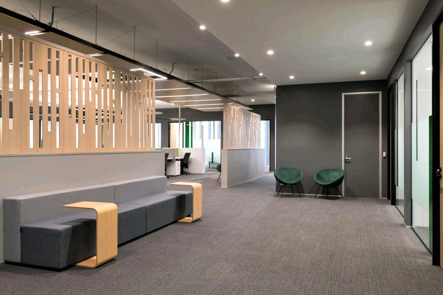 Modern office reception area with gray sofa, green chairs, wooden partitions, and workstations in the background.