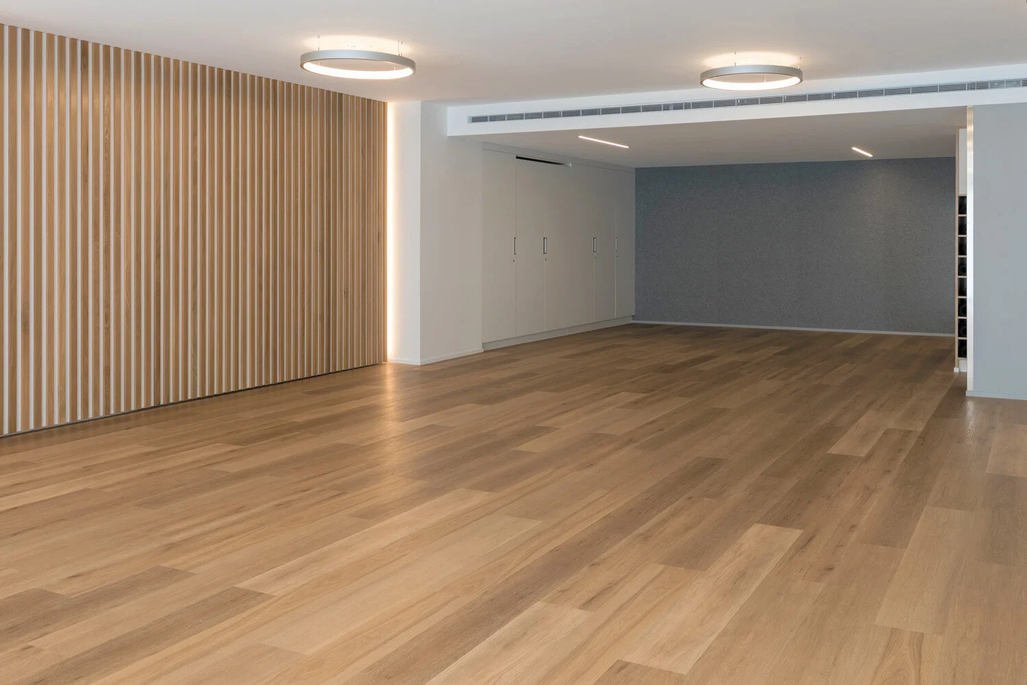 Empty modern room with wooden flooring, white walls, wooden slat accent wall, gray textured wall, and ceiling lights.