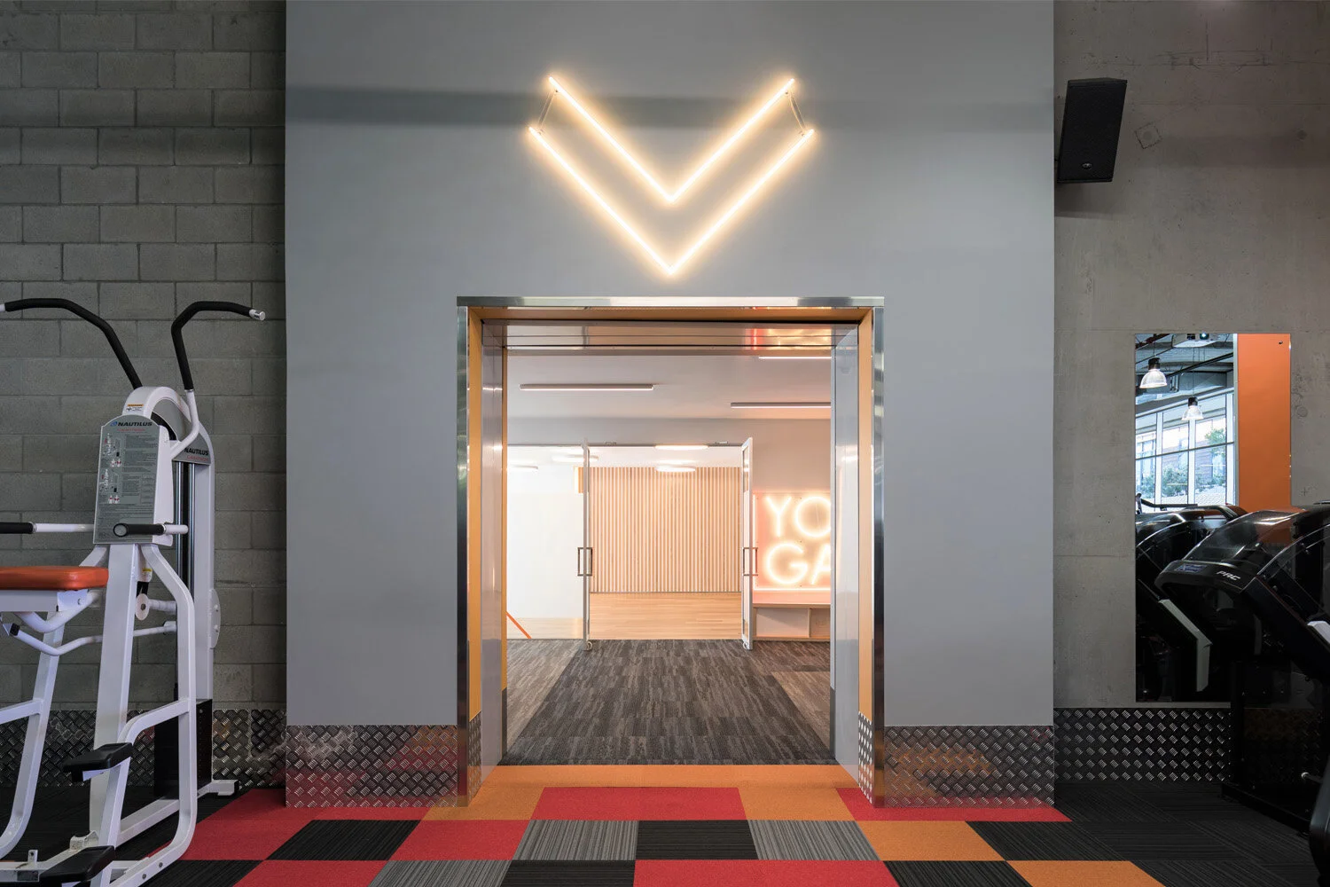 Entrance to a gym with open doors, illuminated neon signs reading "YOGA" and a large LED arrow above the doorway, and gym equipment like a stair climber on the side.
