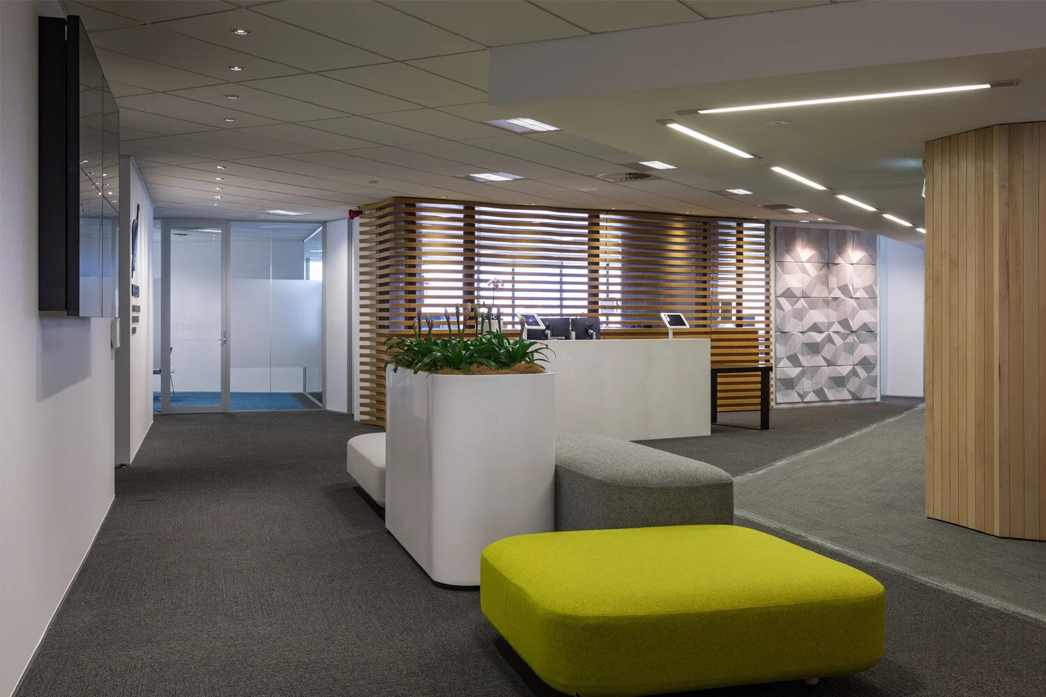 Modern office reception area with a wooden slat partition, a white reception desk, potted plant, colorful seating, and textured wall art.
