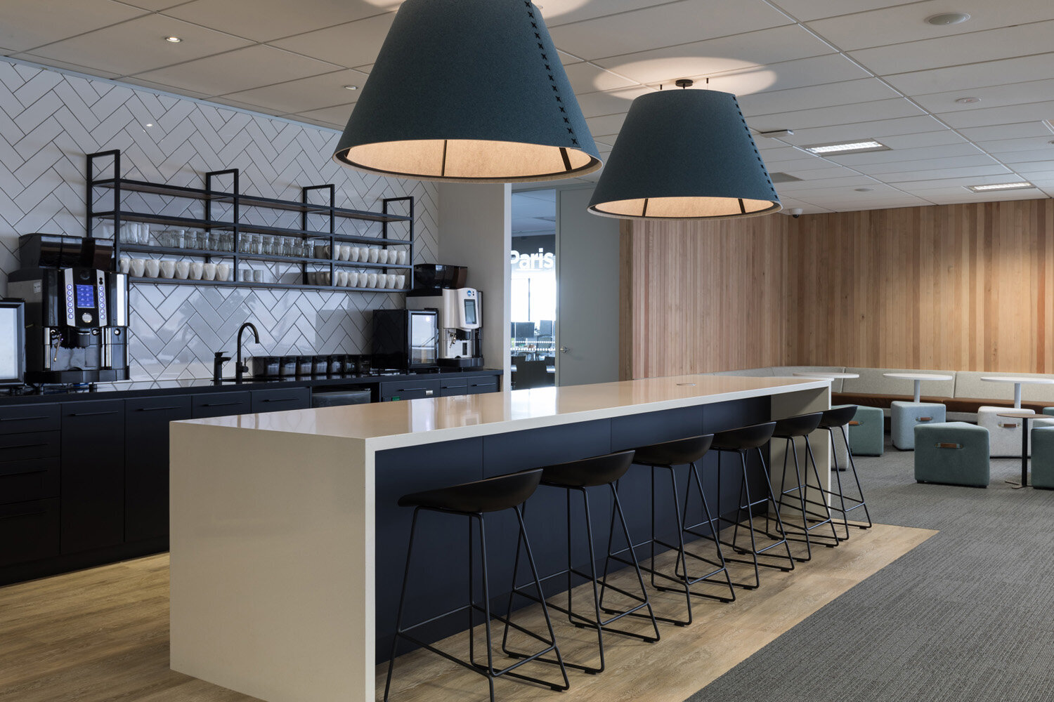 Modern coffee break area with a black and white counter, black stools, coffee machines, and a seating area with stools and chairs.