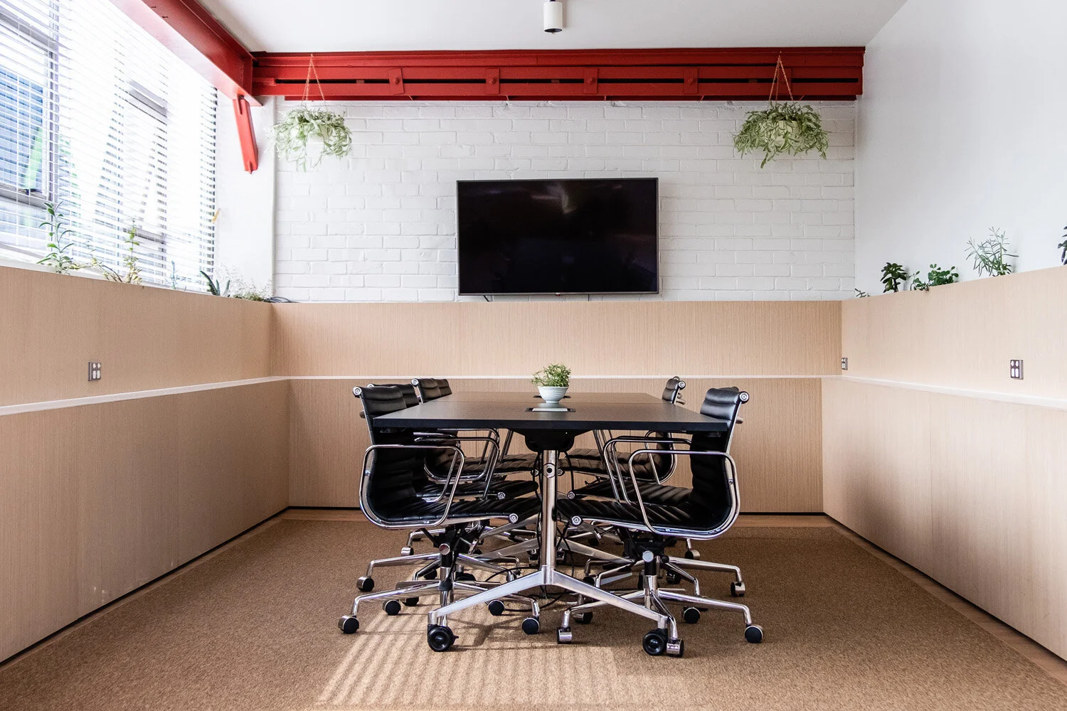 A conference room with a rectangular black table surrounded by six black modern office chairs. There is a potted plant in the center of the table. The room has beige wainscoting and a white brick wall with a large flat-screen TV mounted on it. Natura