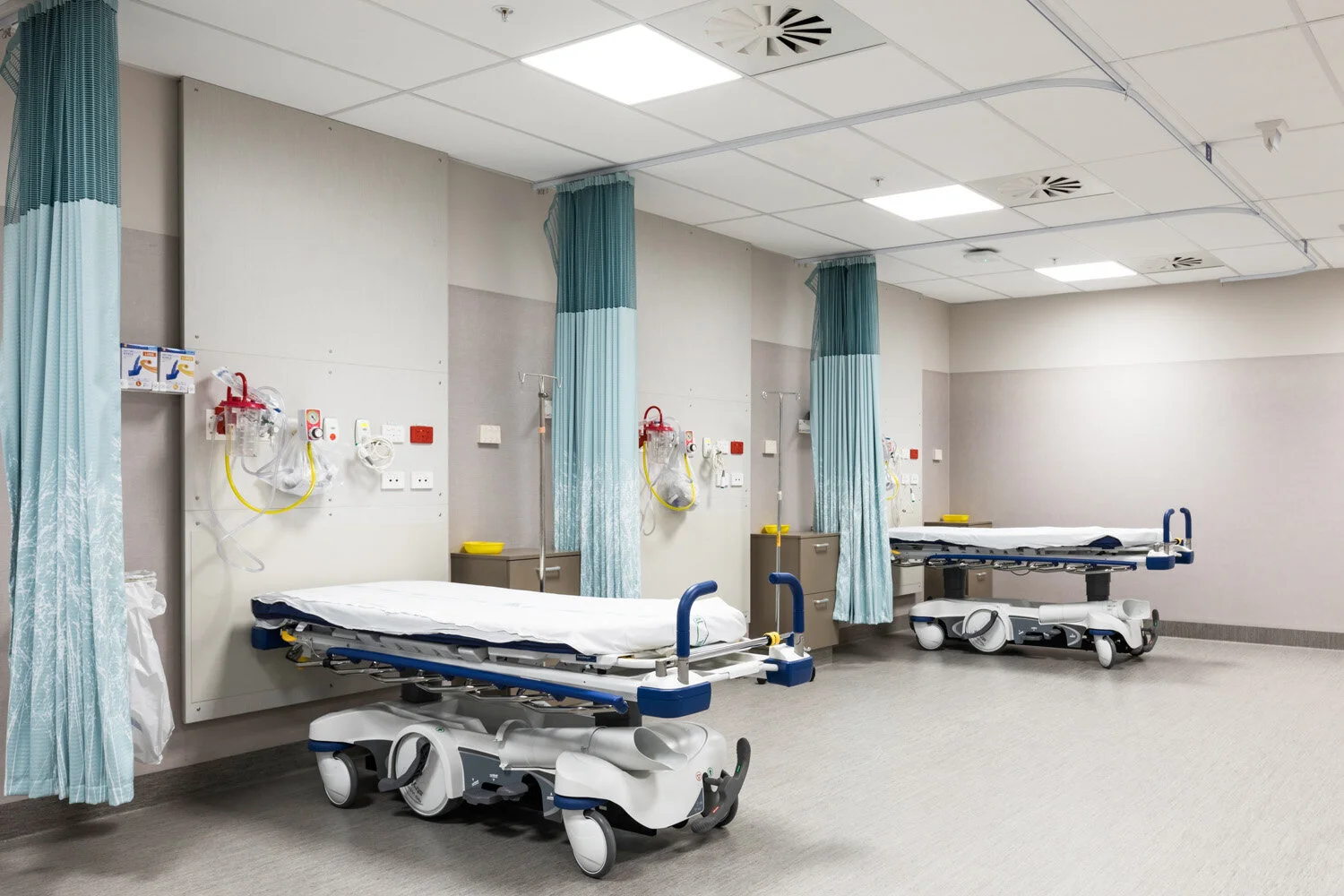 A hospital room with two empty patient beds separated by medical curtains, each bed equipped with medical monitoring and oxygen supplies, in a clean and sterile environment.