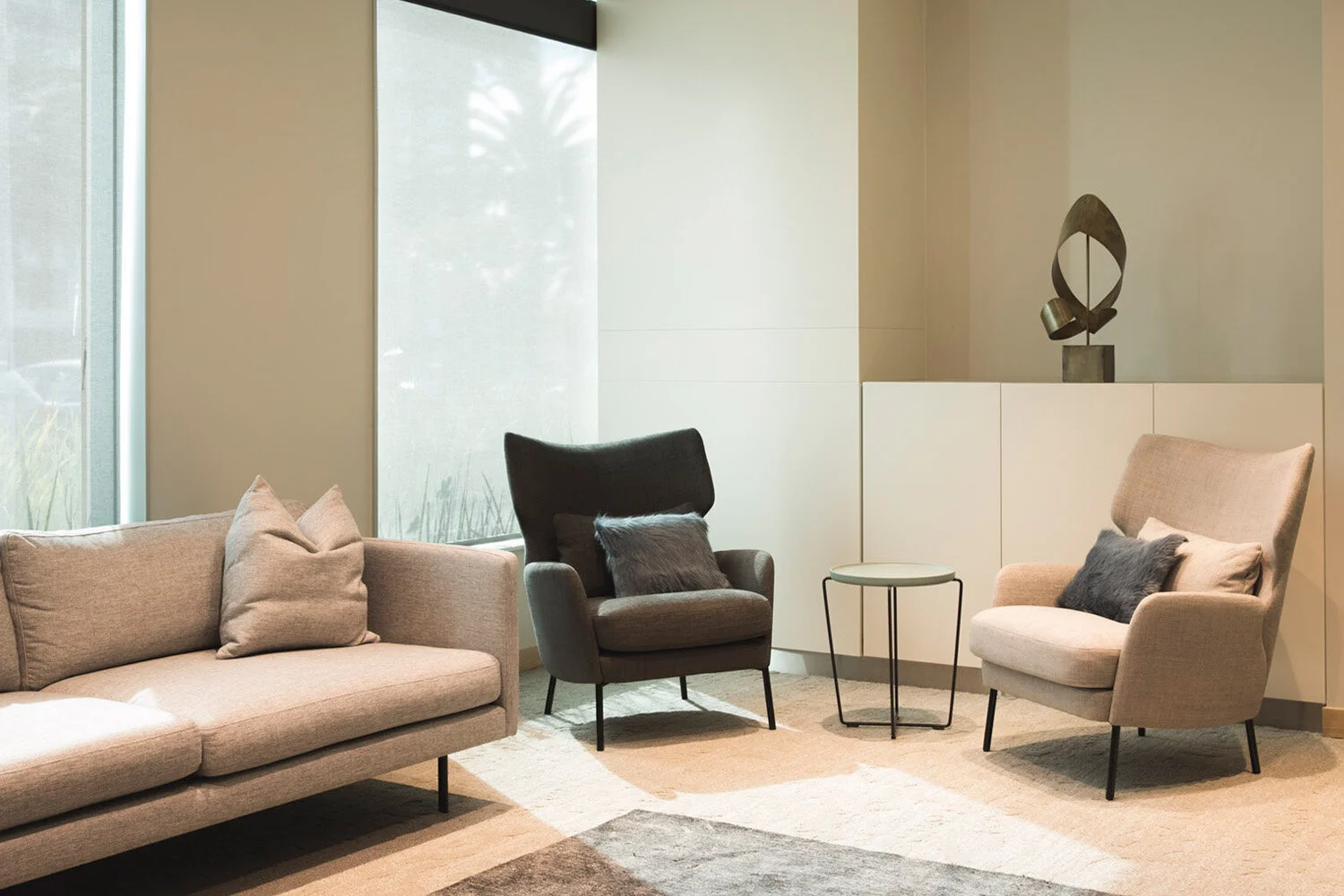 Living room with beige sofa, black armchair, and beige armchair, all with pillows. Small round table between chairs. Window with frosted glass wall. Abstract sculpture on white cabinet.