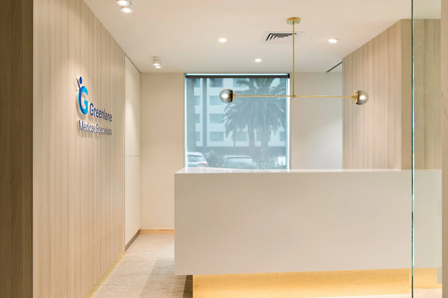 Modern reception area with a white desk, wooden wall paneling, a large window, and a gold and silver decorative light fixture, with the Greenlane Medical Specialists logo on the wall.