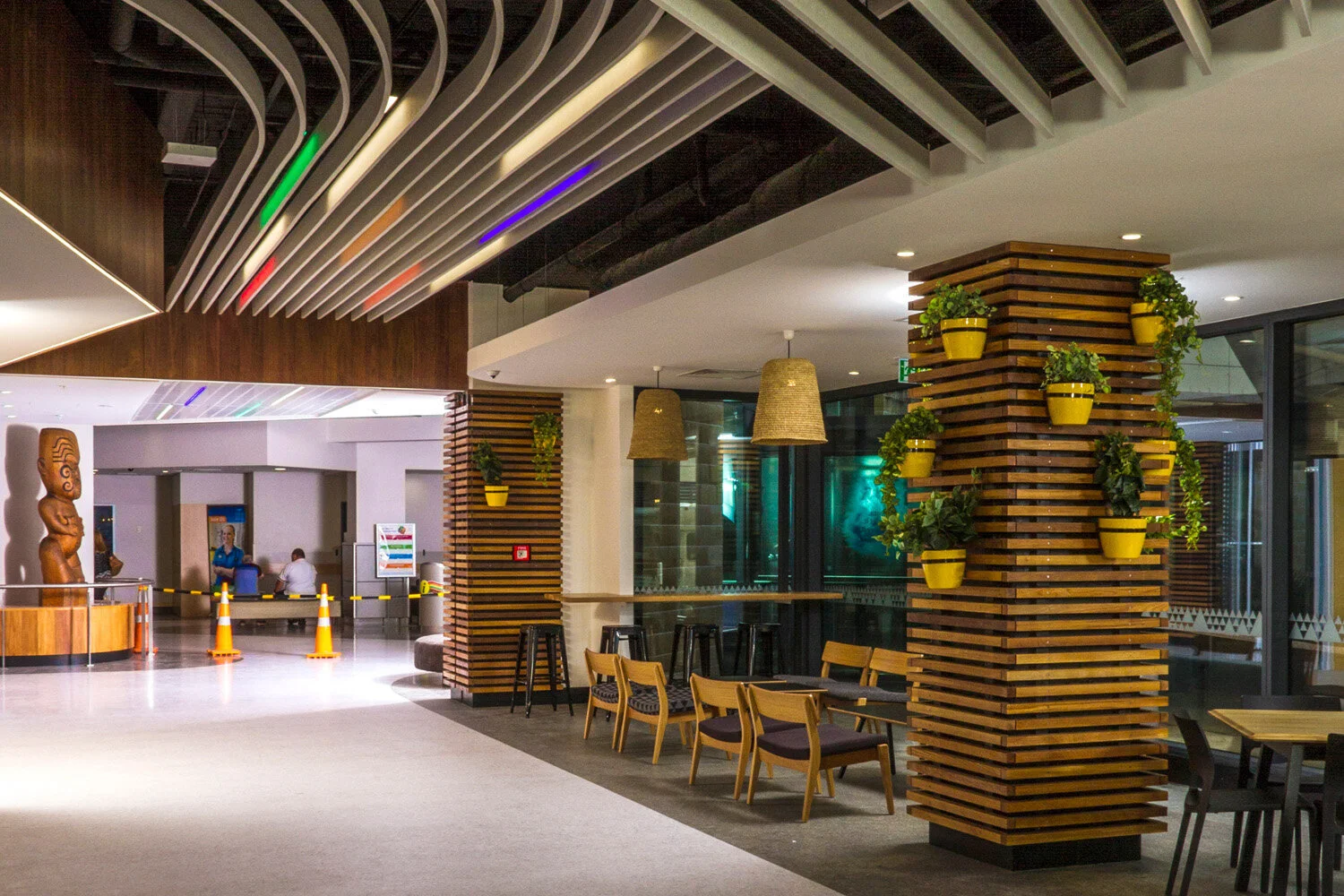 Interior of a modern building lobby or lounge area with wooden and plant decor, seating of chairs and bar stools, hanging woven lamps, and a colorful ceiling design.